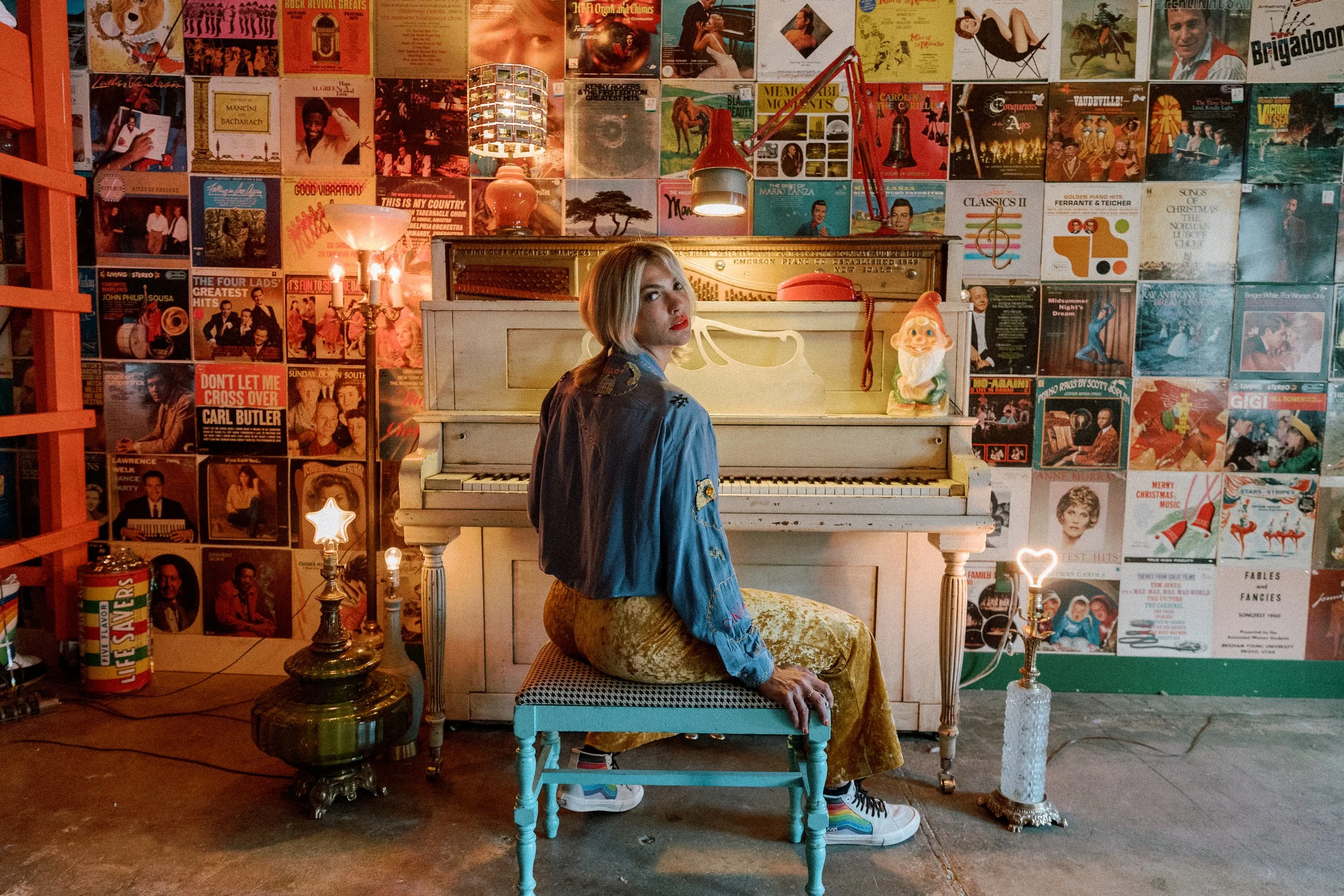 Mindy Gledhill, an indie singer-songwriter, sitting on a small chair in front of a white upright piano. The background wall is covered with colorful album covers. There are lamps, one with a star and another with a heart, beside her.