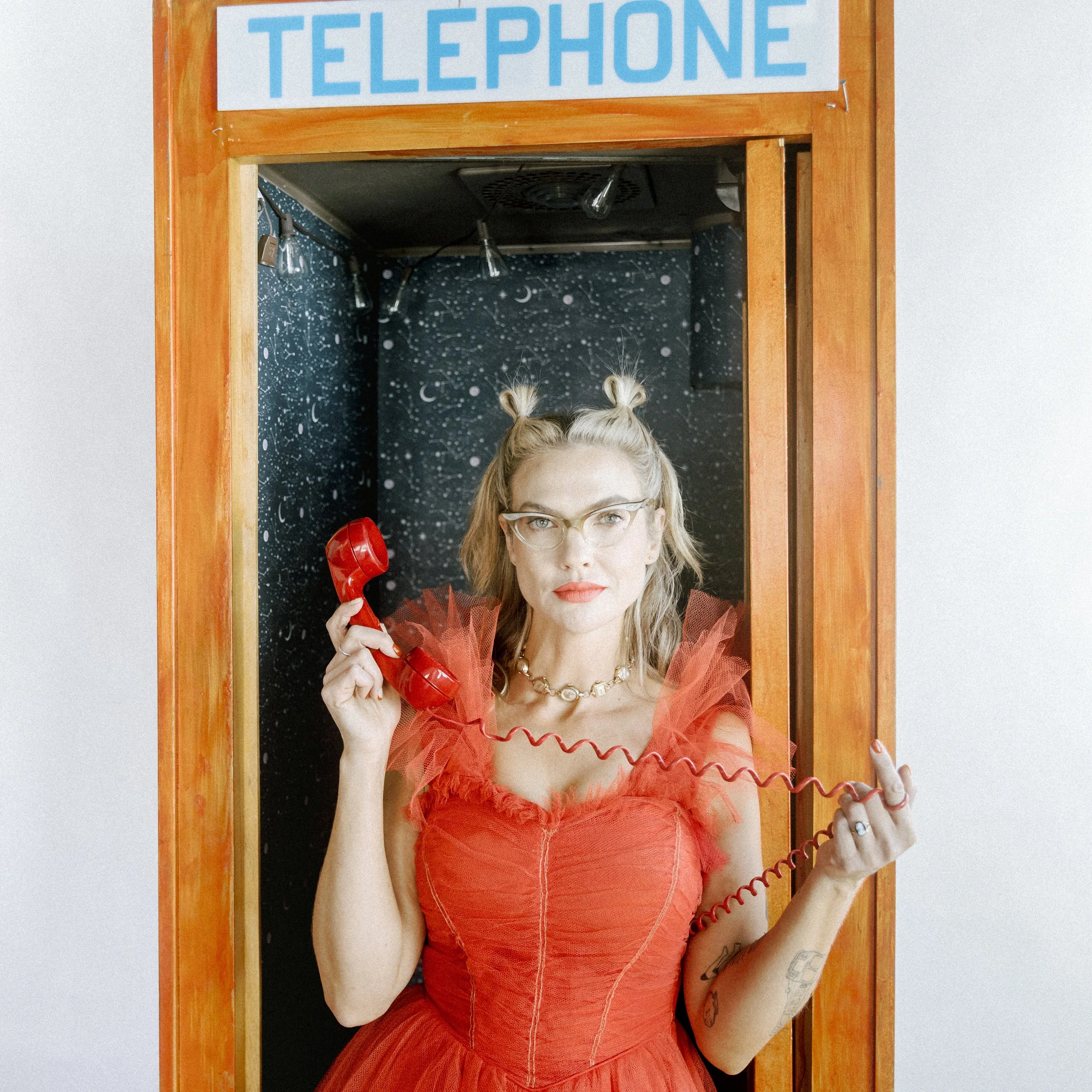 Mindy Gledhill, an indie singer-songwriter, in a red dress and glasses standing inside a vintage orange phone booth, holding a red rotary phone receiver to her ear, with a galaxy-themed wallpaper background.