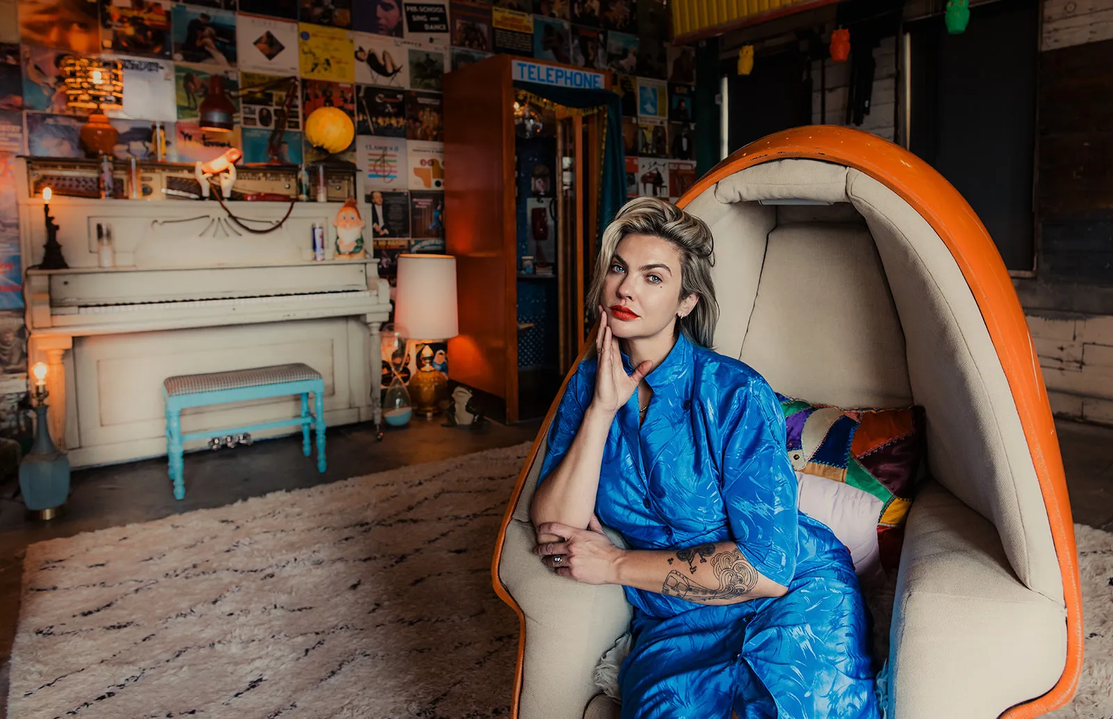 A woman with blonde hair and red lipstick sitting inside an orange, pod-like chair in a decorated room with a piano, wall art, and a lamp.