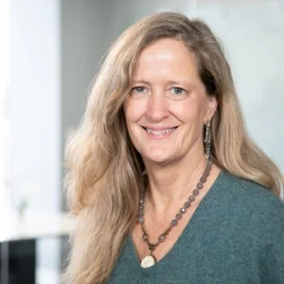 A woman with long, wavy blonde hair smiling, wearing a teal top, earrings, and a beaded necklace, standing indoors with a blurred background.