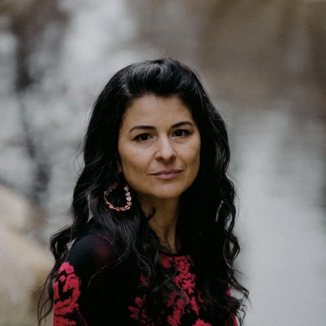 A woman with long, dark, curly hair wearing a black and red patterned top and a large pink hoop earring. She is outdoors with a blurred, natural background.