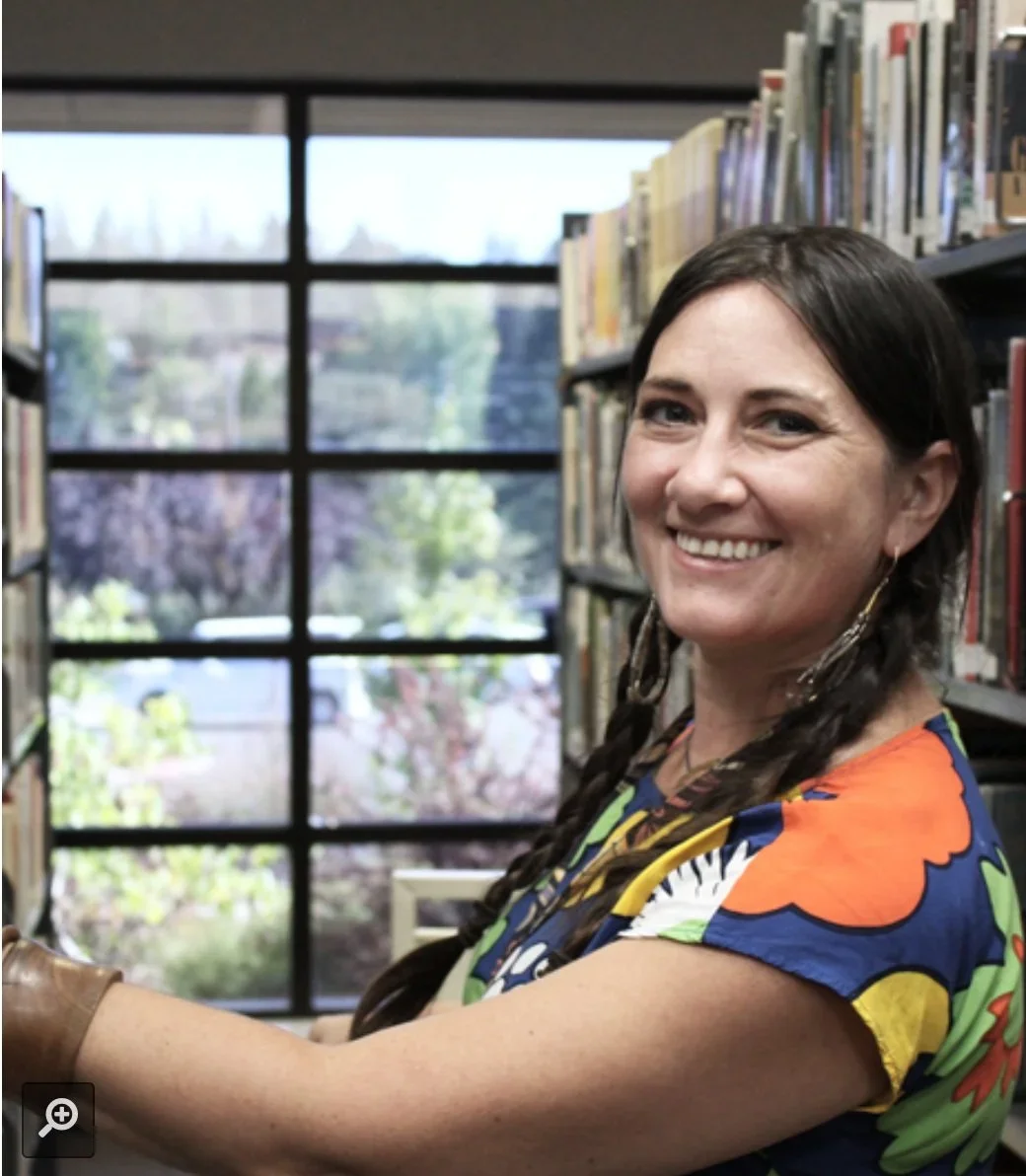 A woman with dark brown hair in braids, smiling in a library with bookshelves and a window showing trees outside.