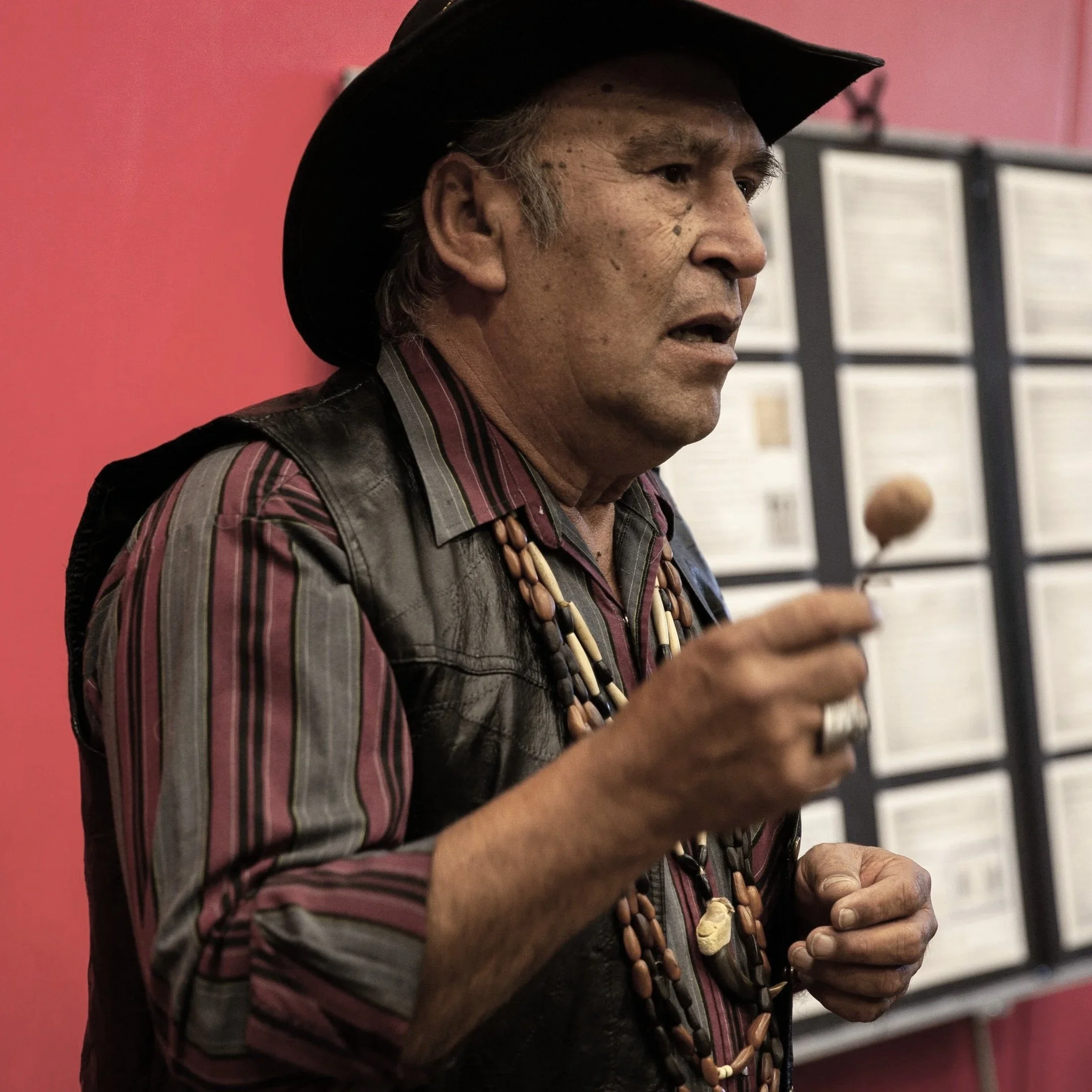 An older man with a dark hat and jewelry, wearing a patterned shirt with a vest, holding a small object, standing in front of a pink wall with framed documents.