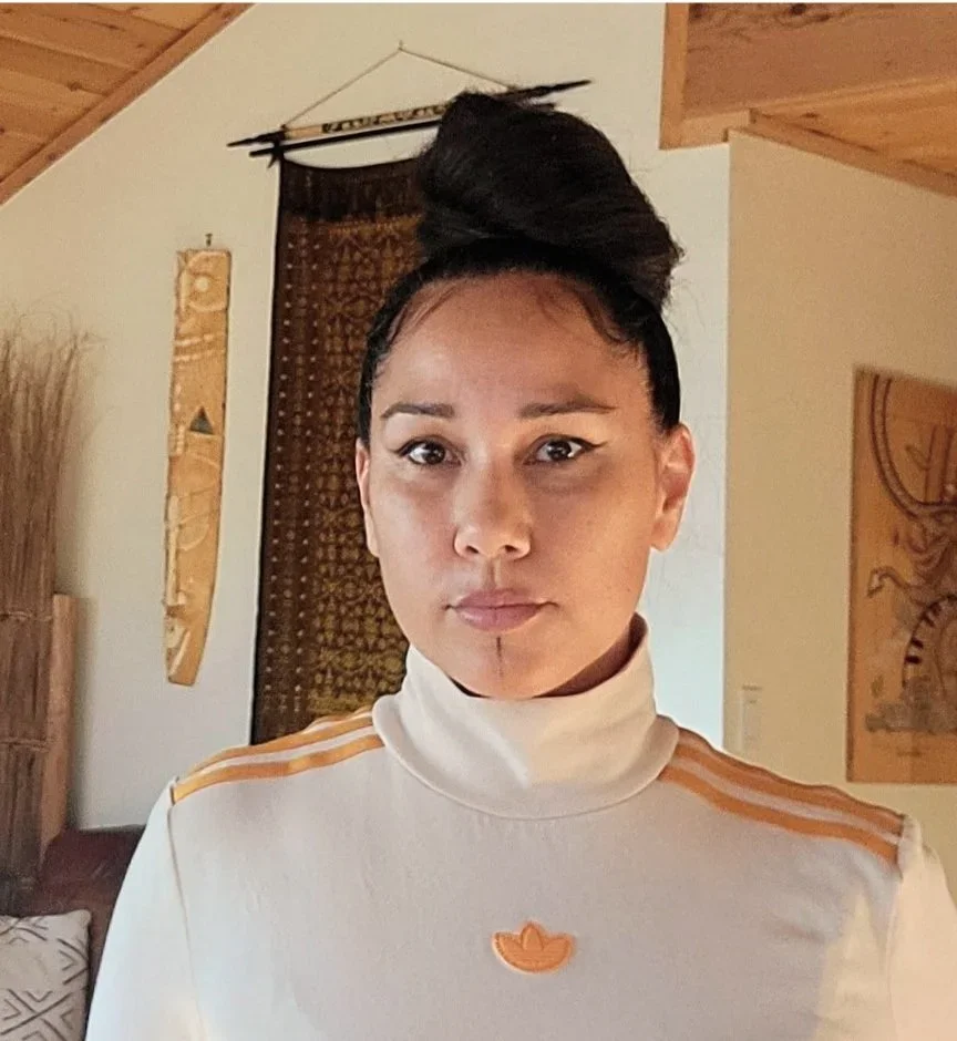 A woman with dark hair styled in a high, voluminous top knot, wearing a white turtleneck with gold accents on the shoulders, standing in a room with tribal and nature decor.