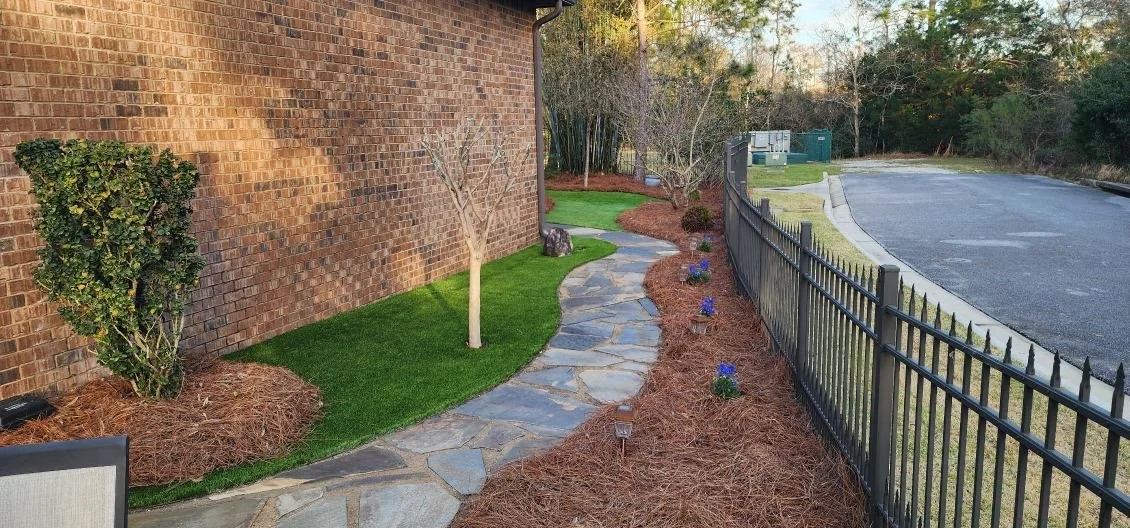Stone pathway along a landscaped yard with small trees, bushes, and blue flowers, bordered by mulch, next to a brick building and a black metal fence.