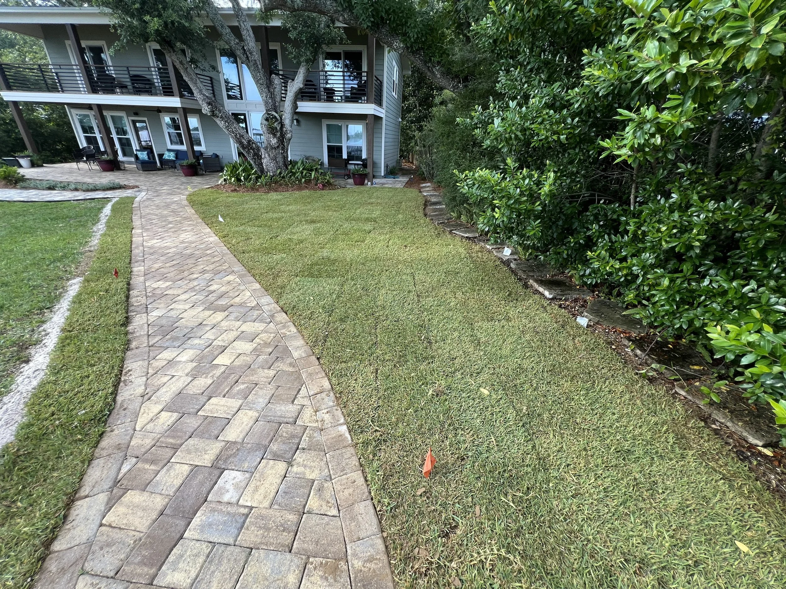 A curved brick pathway leads towards a gray house with a porch, surrounded by a well-maintained lawn and lush green bushes on the right side.