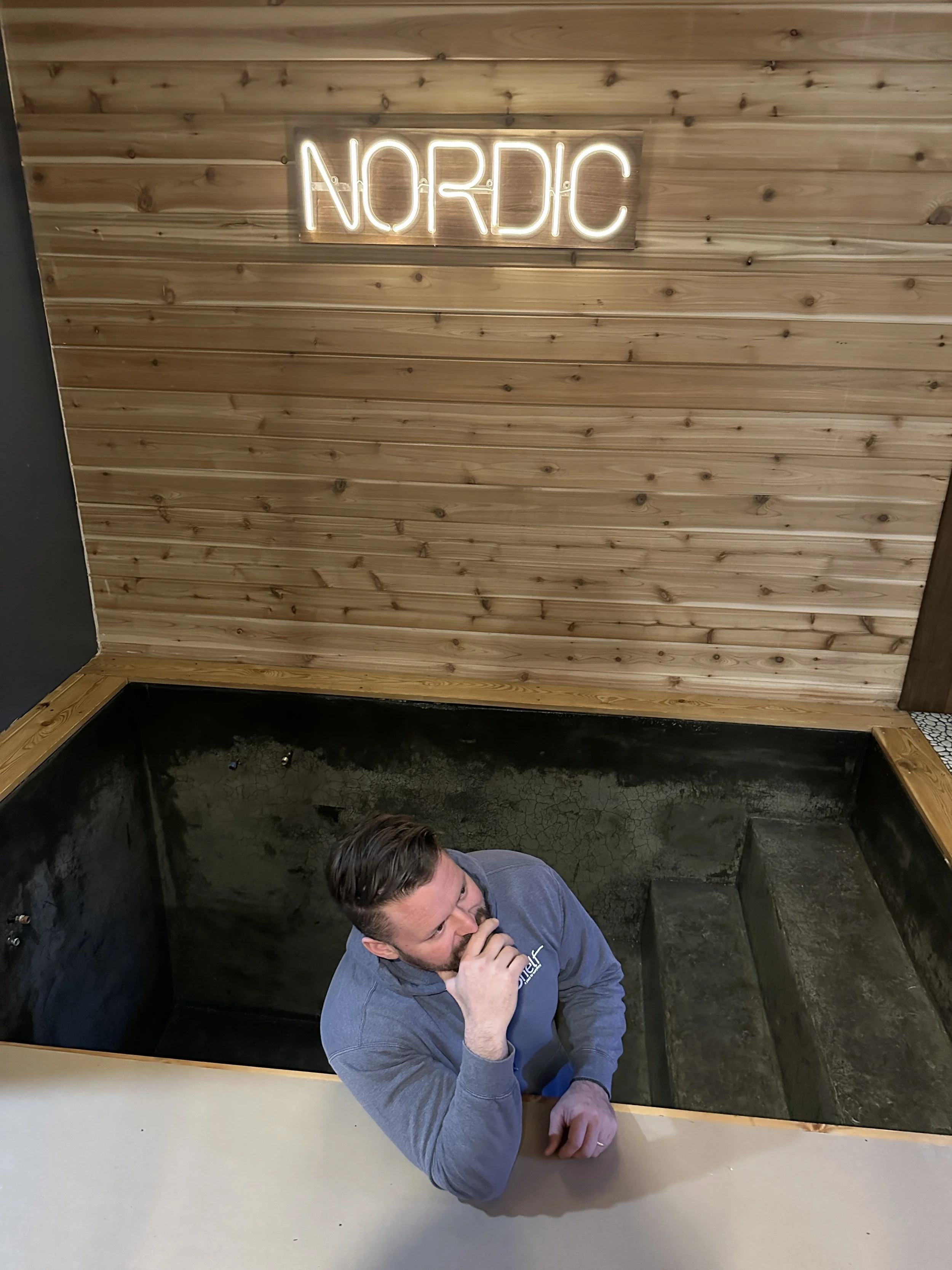 Nordic on Nine owner sitting in cold plunge pool beneath the Nordic neon sign at the New Windsor NY wellness spa