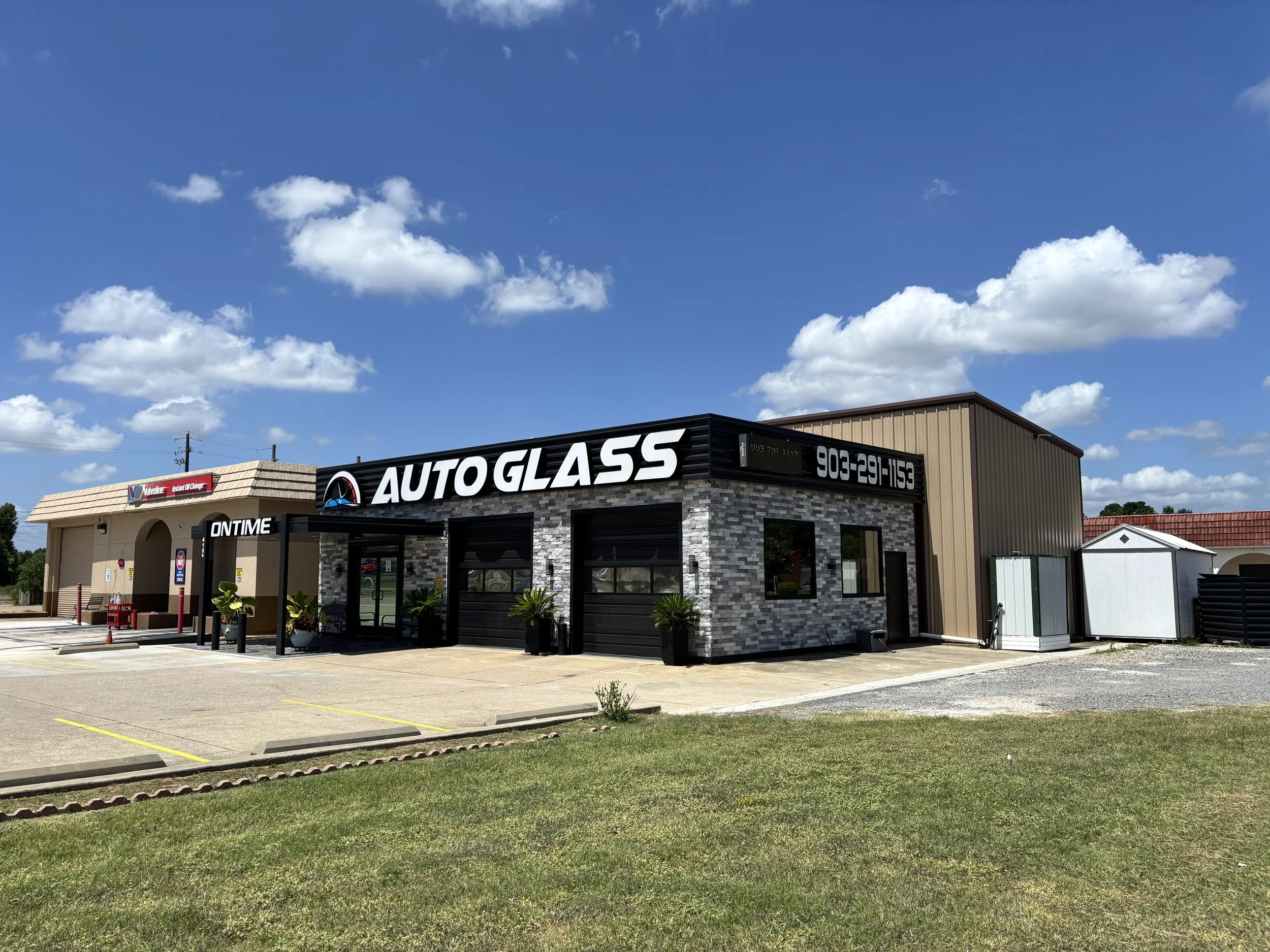 A building with a large sign that reads 'AUTO GLASS' and a phone number, with a smaller adjacent building on the left and some potted plants near the entrance, under a blue sky with clouds.
