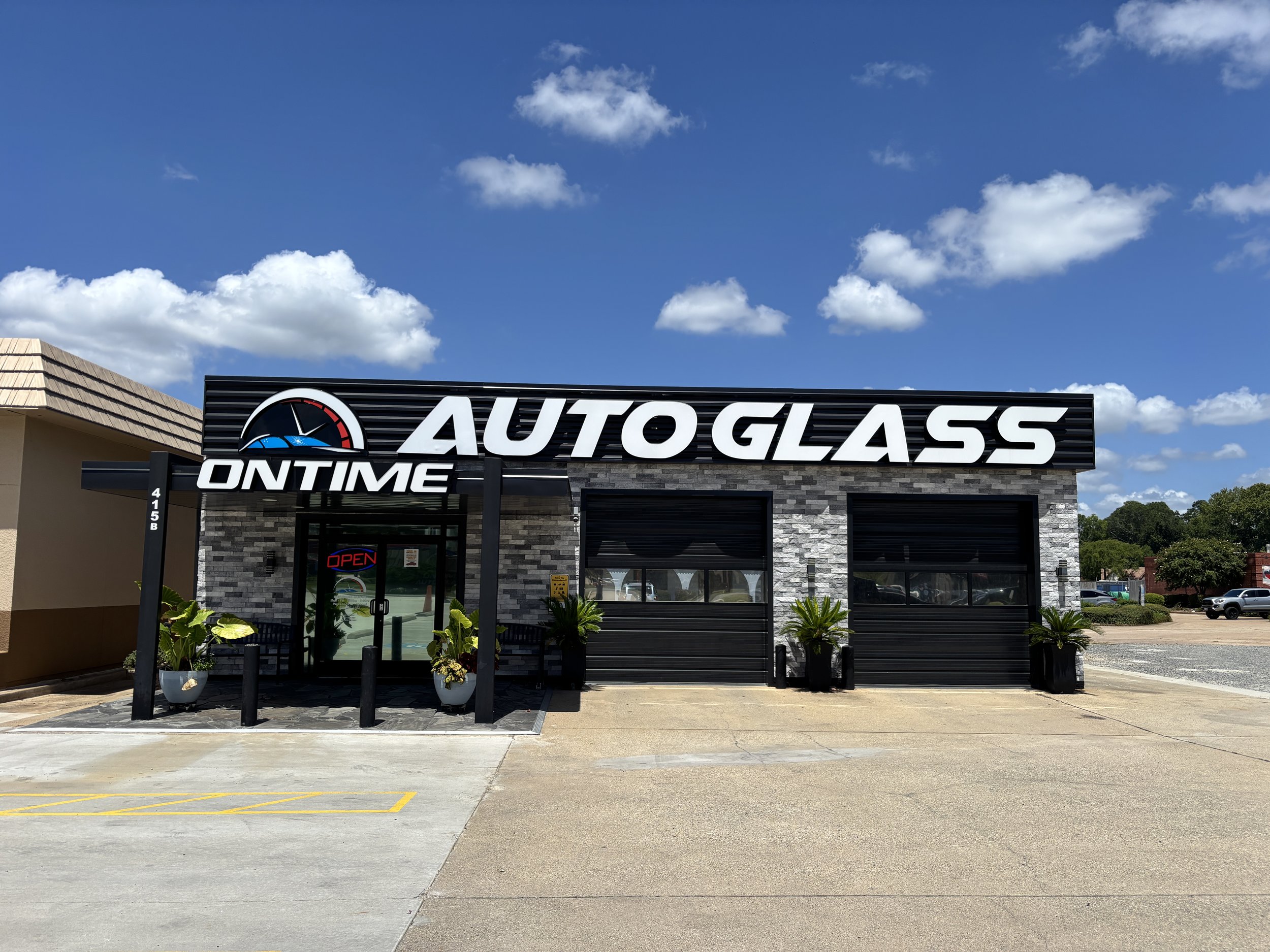 Front view of an auto glass repair business called 'AUTO GLASS ON TIME' with black and grey exterior, two garage doors, a glass entrance door with an 'OPEN' sign, and potted plants, under a blue sky with scattered clouds.