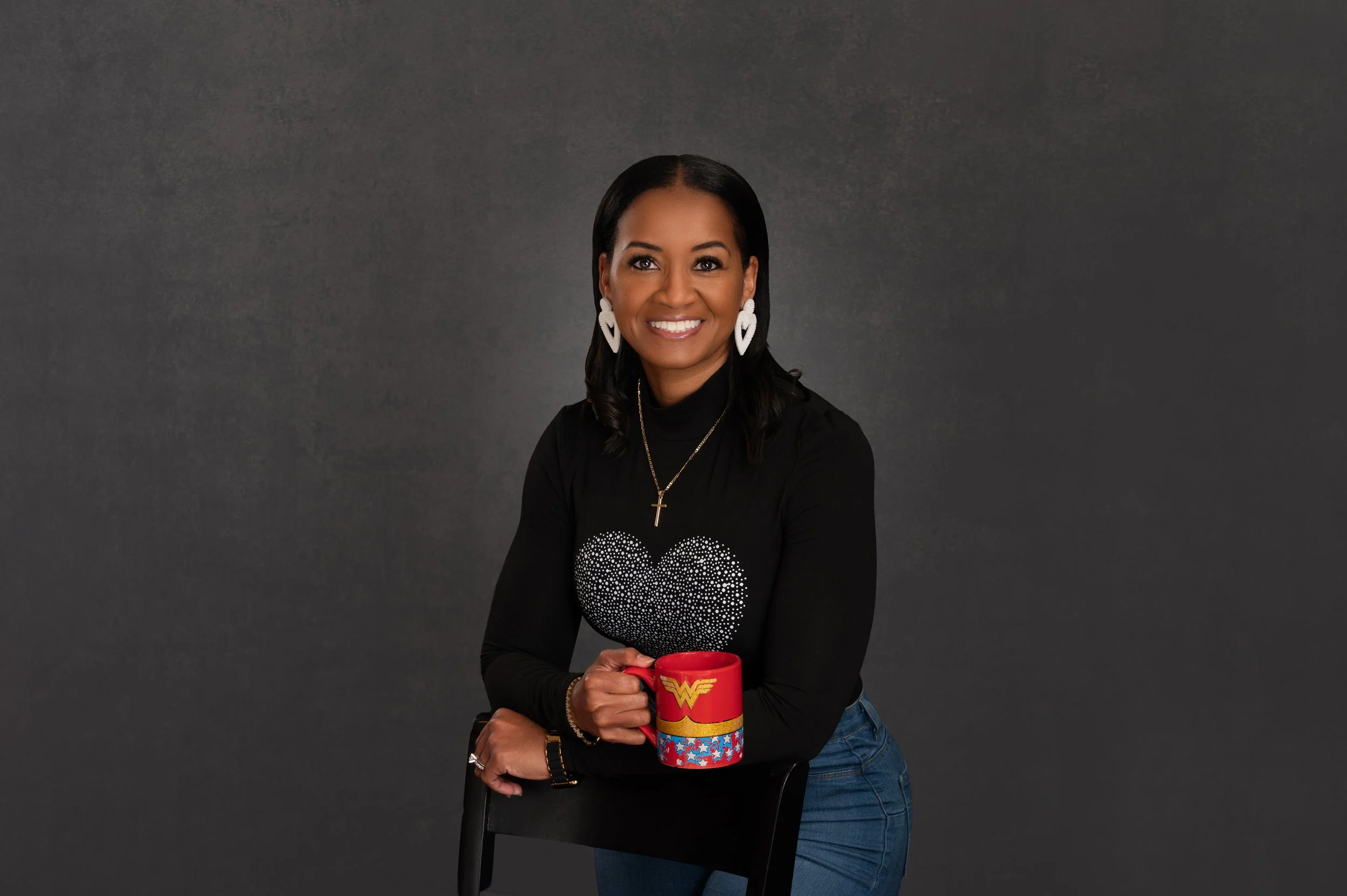A smiling woman with dark hair and earrings, wearing a black top with a heart design and a Wonder Woman mug, sitting on a black chair against a dark gray background.