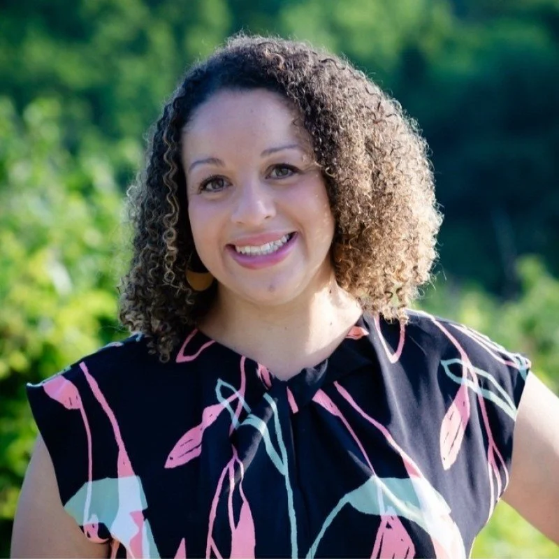Smiling woman with curly hair outdoors, wearing a black top with pink and white abstract patterns.