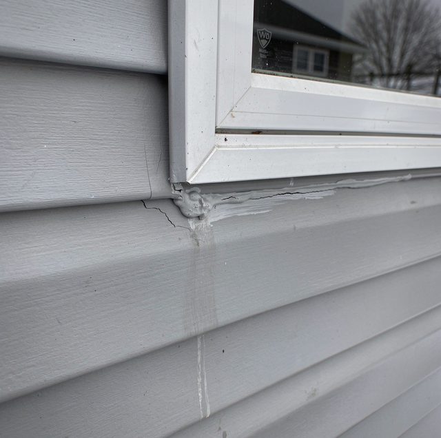White vinyl siding and window frame on exterior wall with reflection of house and bare tree visible in glass.