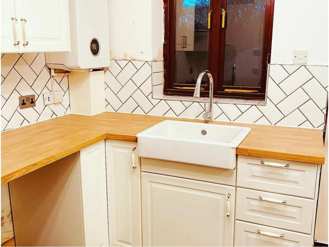 Kitchen with white cabinets, a wooden countertop, a white farmhouse sink, a window above the sink, and a tiled backsplash with a herringbone pattern.