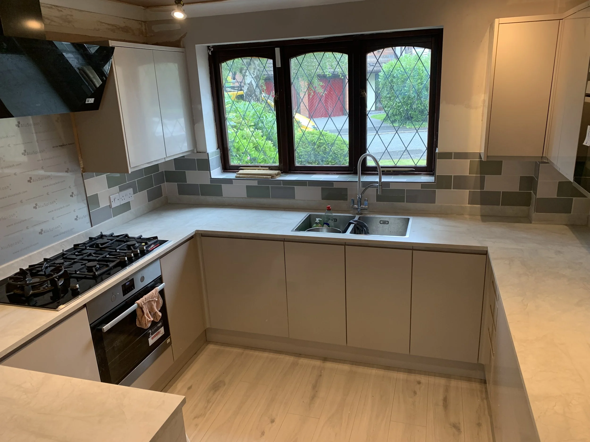 Modern kitchen with beige cabinets, a granite countertop, a gas stove, a built-in oven, a stainless steel sink under a window, and a gray backsplash with rectangular tiles. A window with a decorative grille shows a garden outside.