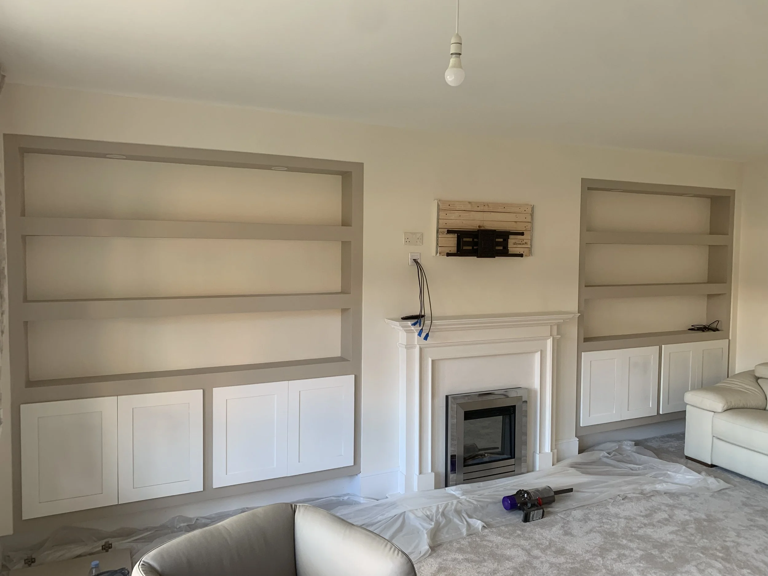 Living room under renovation with built-in bookshelves, a fireplace, and a white couch. There are construction tools and protective sheets on the carpeted floor.