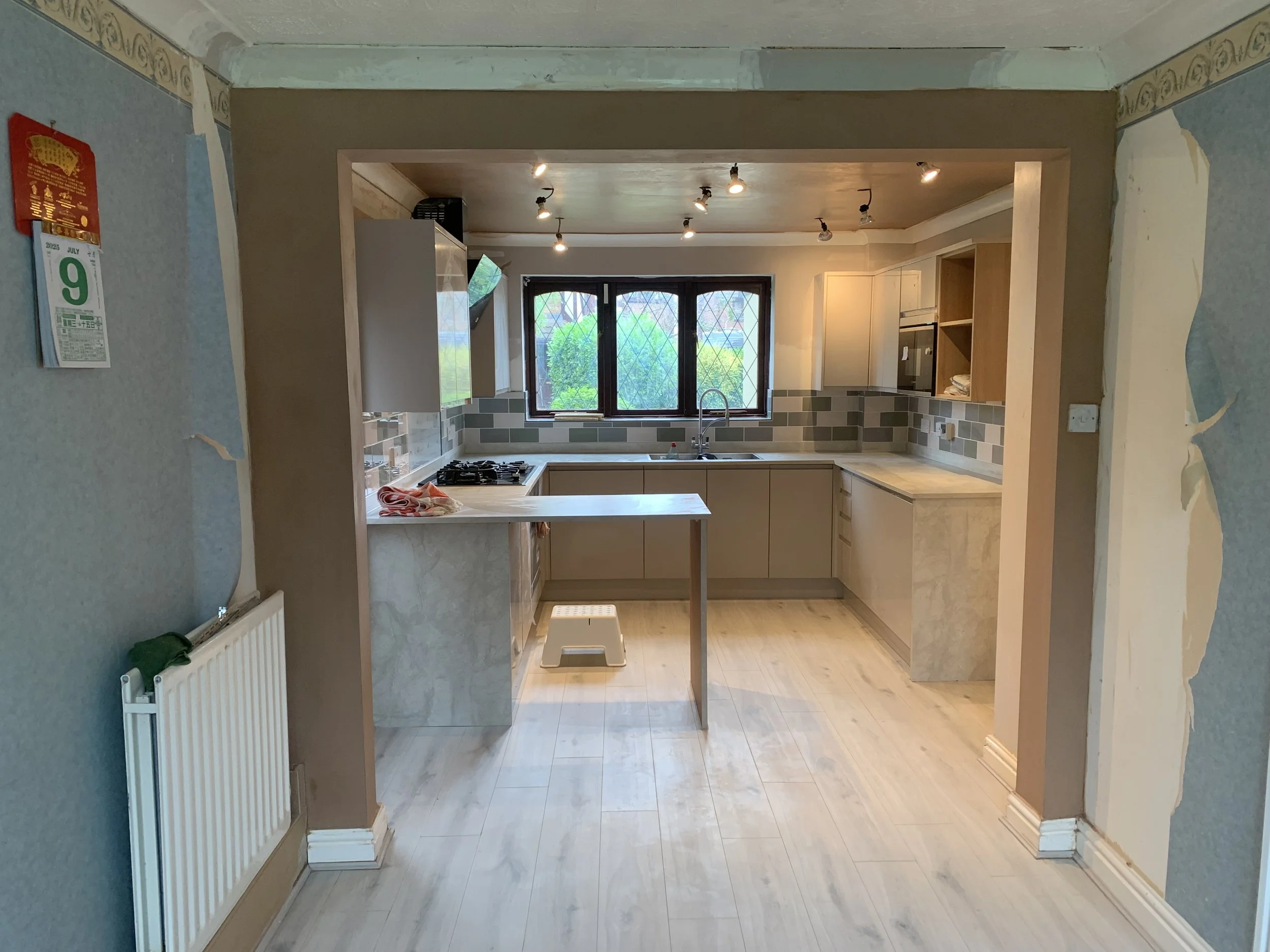 The image depicts a kitchen under renovation with partially installed cabinets, a small counter extension, a window above the sink, a stove, and a tiled backsplash. The walls are partly painted, and there are ceiling lights and a stool on the floor.