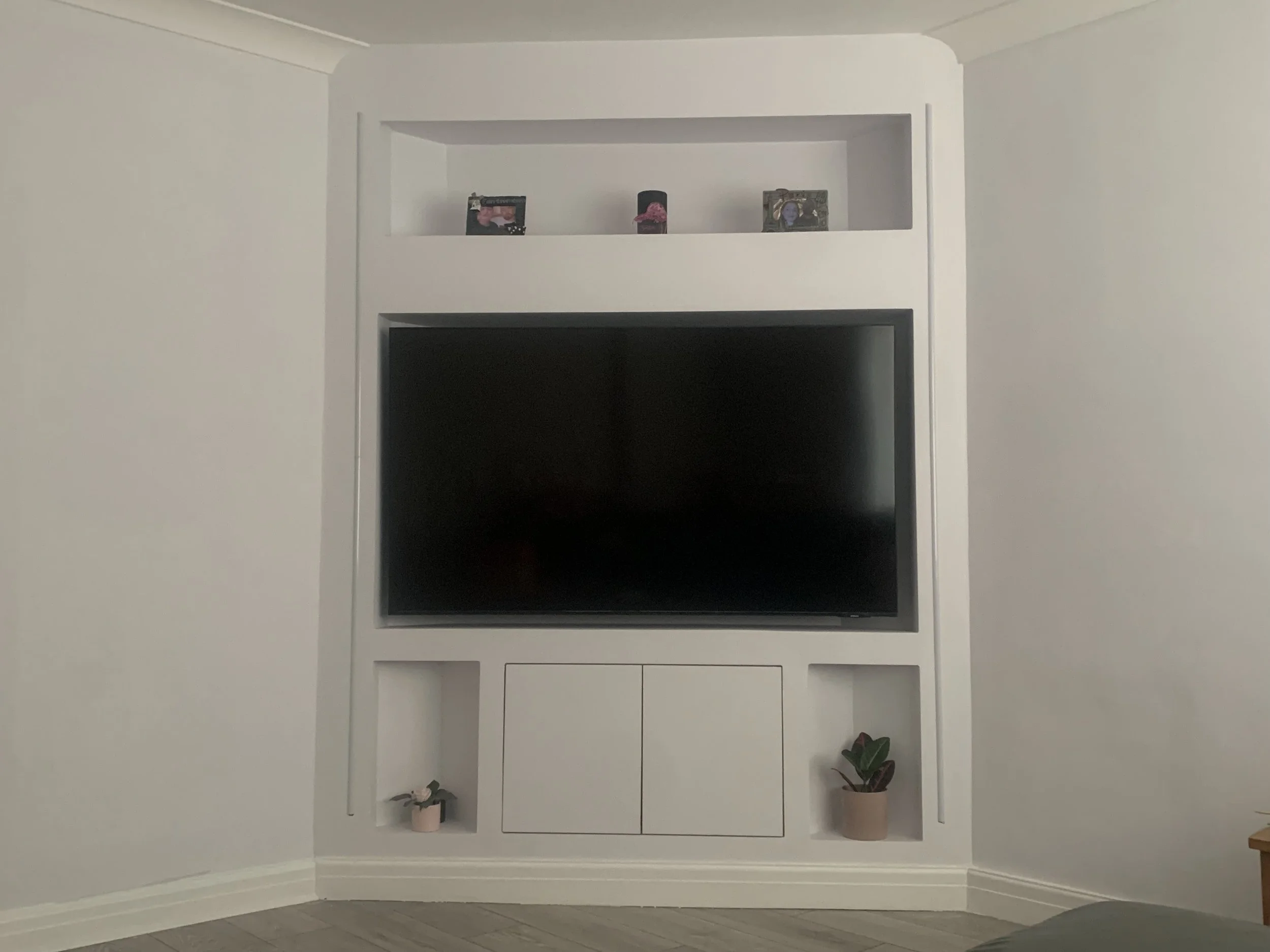 White built-in entertainment center with a large flat-screen TV, decorative items, framed photographs on the top shelf, and potted plants on the bottom shelves, against a beige wall.