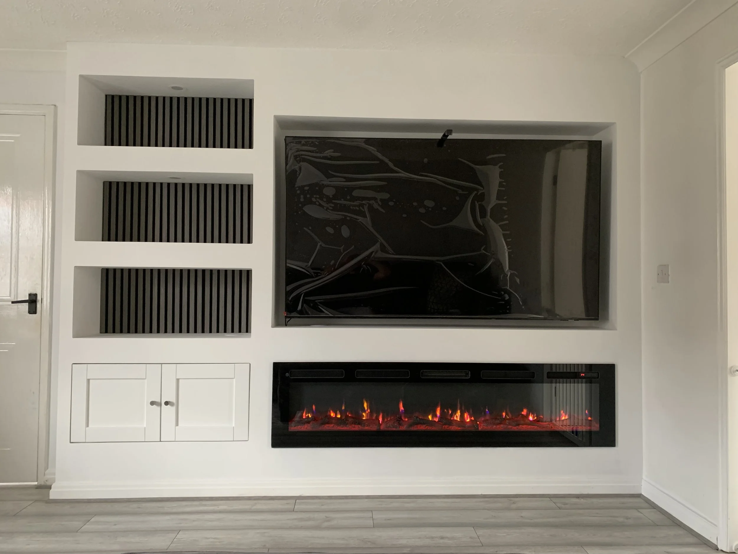 Wall-mounted electric fireplace with orange and red flames, built into a white wall below a large flat-screen TV, surrounded by white shelving and cabinets, with wood-look flooring.