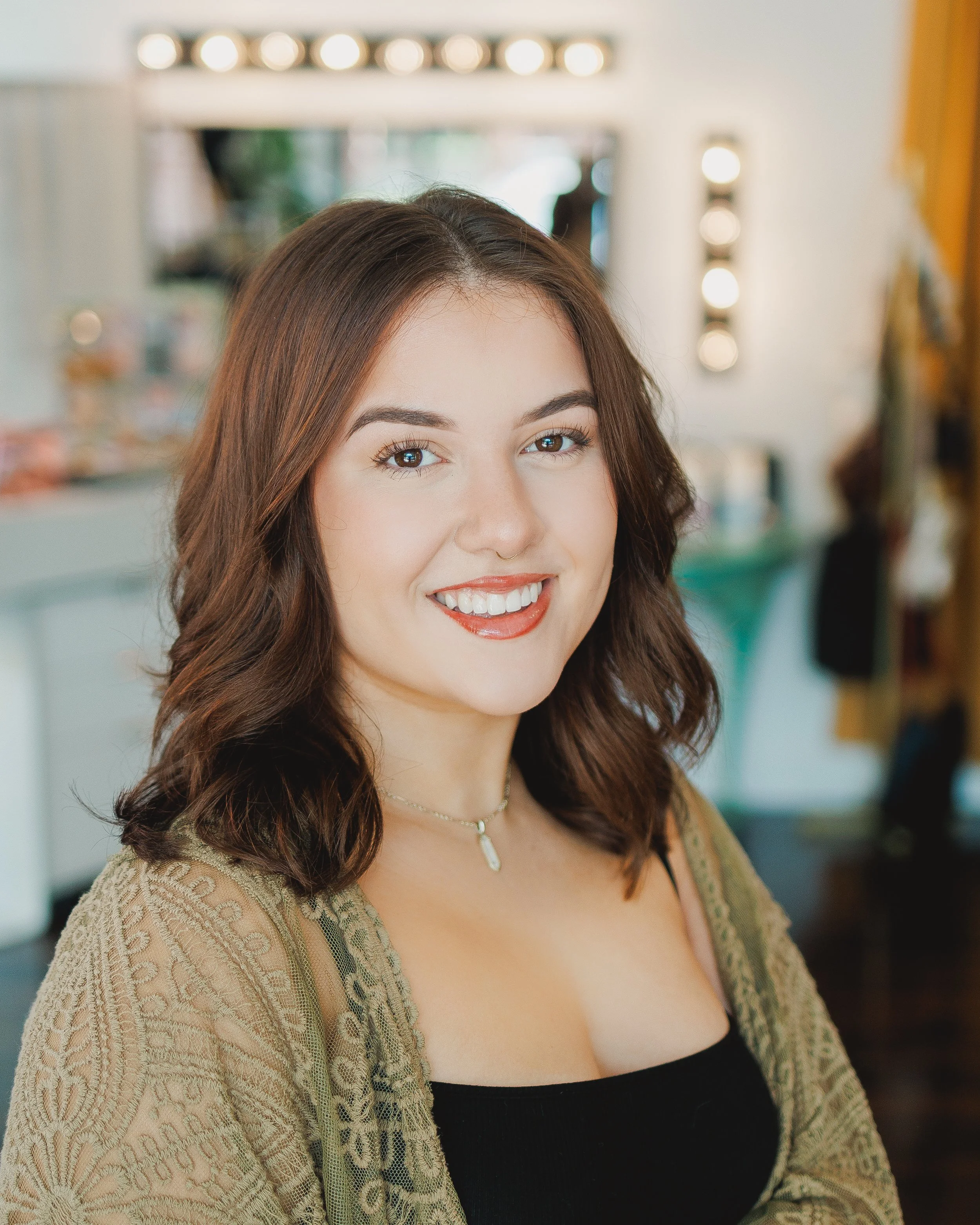 A smiling woman with long brown hair and a nose ring, wearing a black top and a beige lace cardigan, indoors with a mirror and lights in the background.
