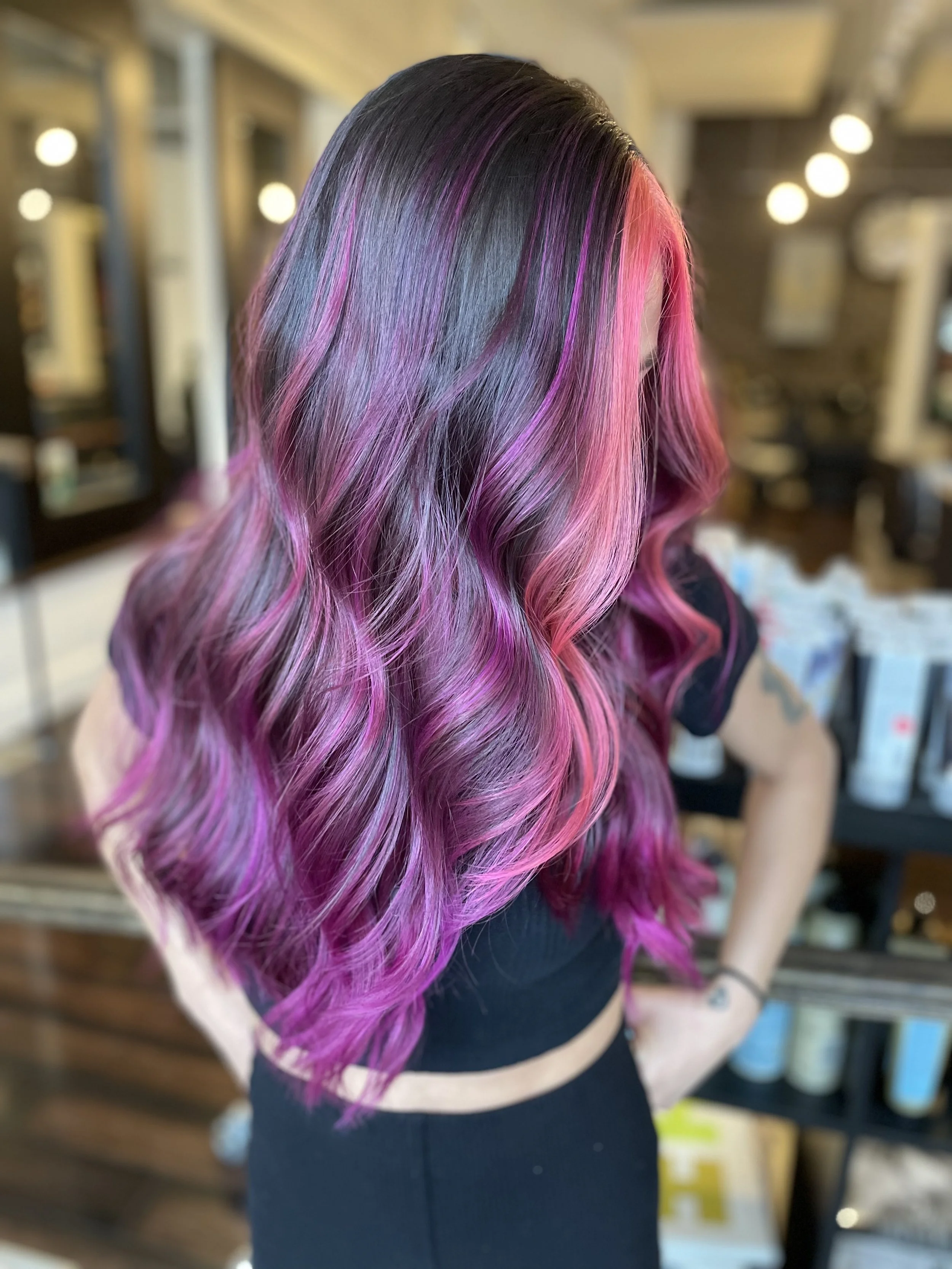 Person with long, wavy hair dyed in shades of pink and purple, standing in a salon.