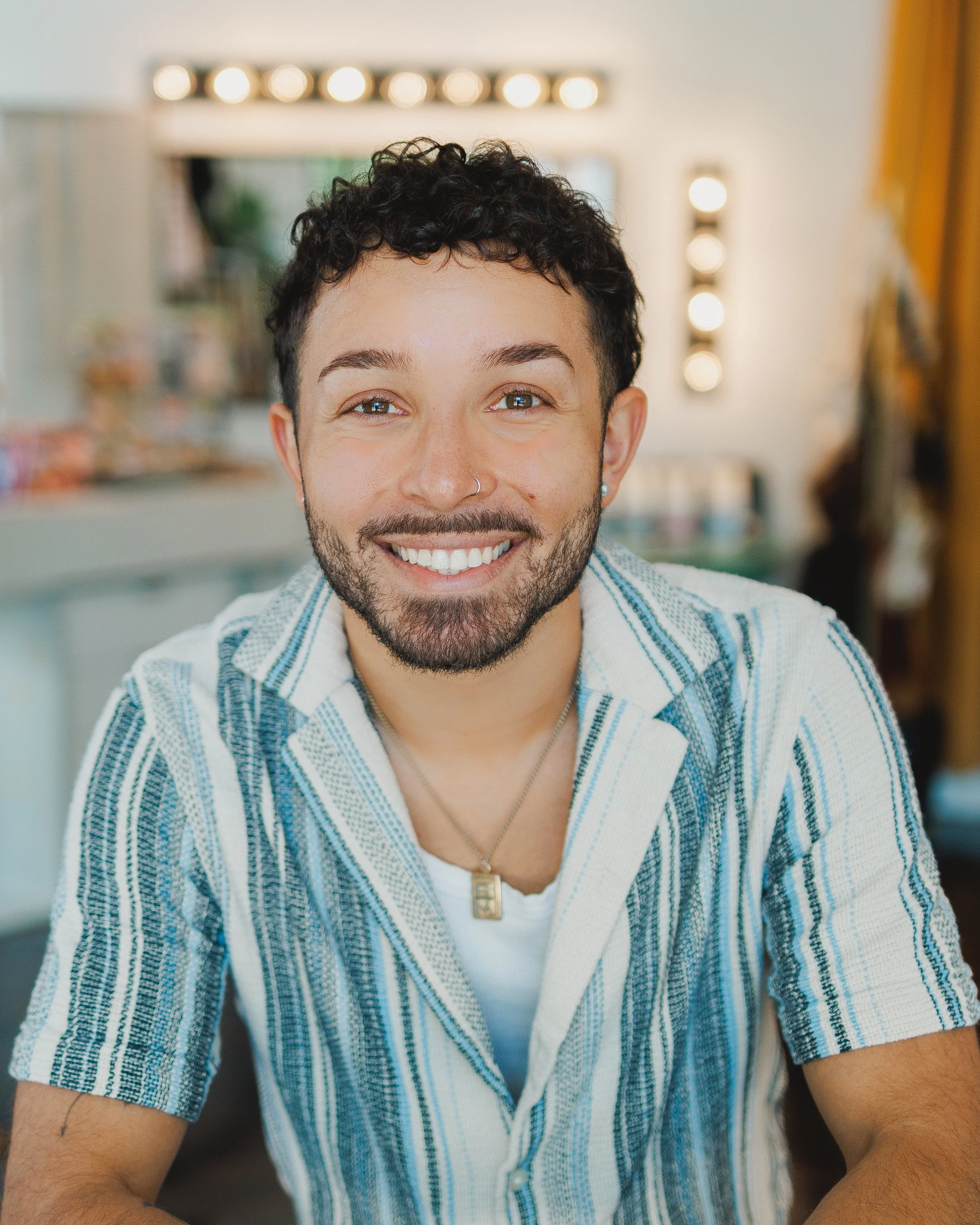 A smiling person with short curly dark hair, a nose ring, and a beard, wearing a striped blue and white shirt, with a blurred background of a room with warm lighting.