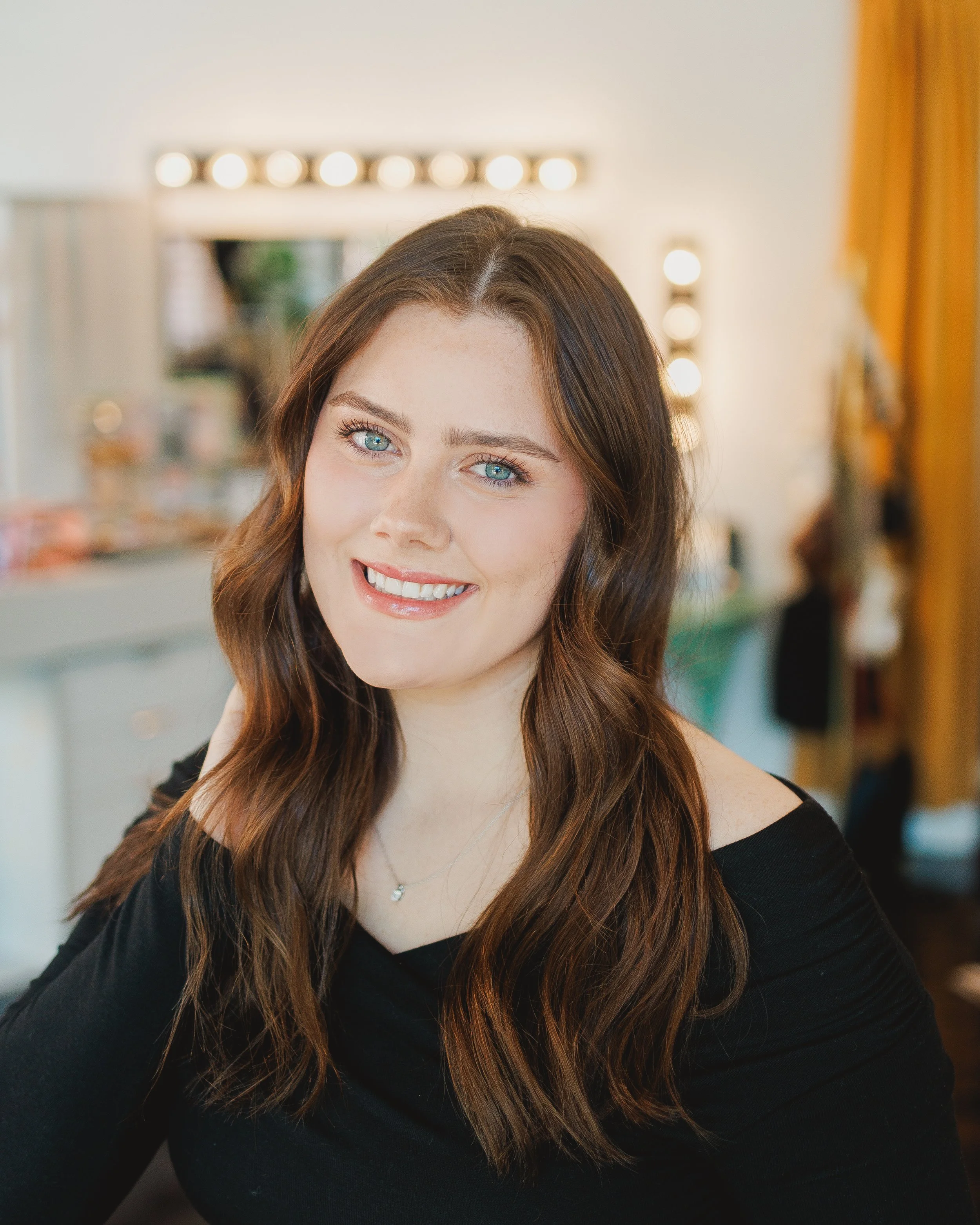 Close-up of a smiling woman with brown hair and blue eyes, wearing a black off-shoulder top, in a brightly lit room with blurred background.