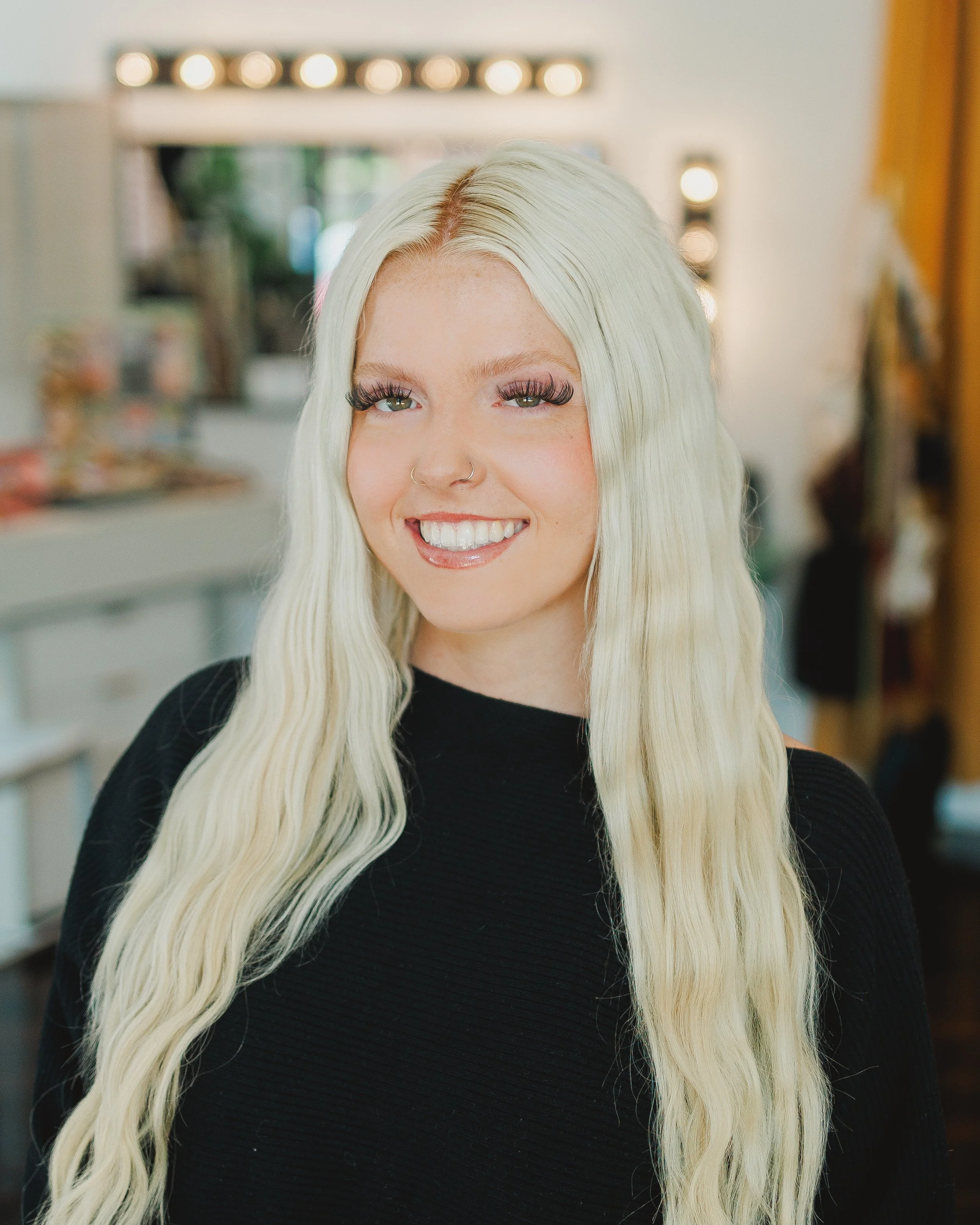 A young woman with platinum blonde hair, green eyes, and long wavy hair, wearing a black top, smiling with makeup including false eyelashes, in a room with blurred background and warm lighting.