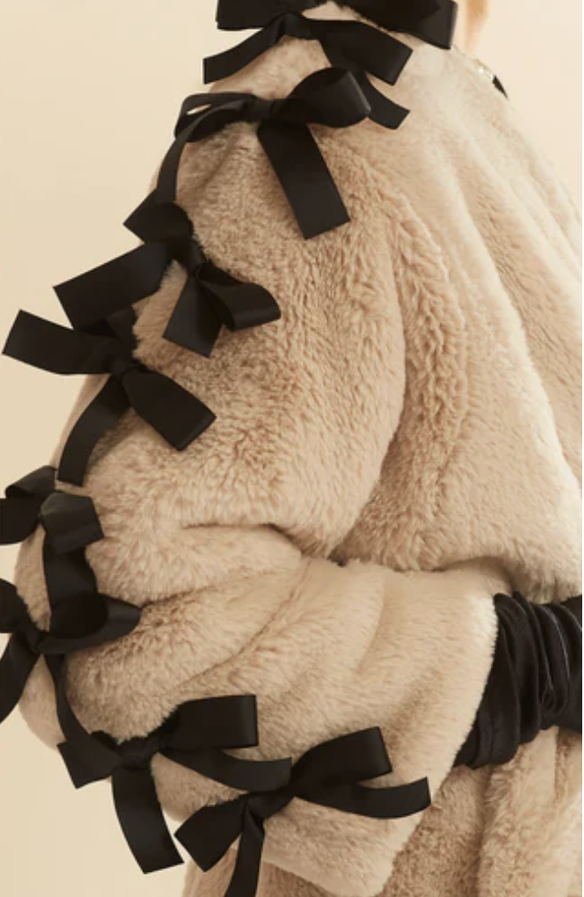 Close-up of person wearing a beige, fluffy coat with black ribbon bows along the shoulder and sleeve.