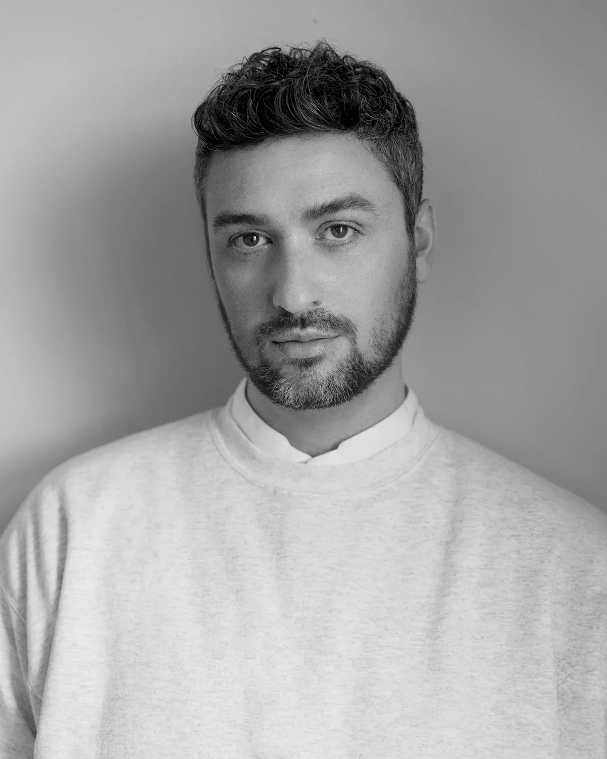 Black and white portrait of a man with short curly hair, beard, wearing a light-colored crewneck sweater over a collared shirt, standing against a plain background.