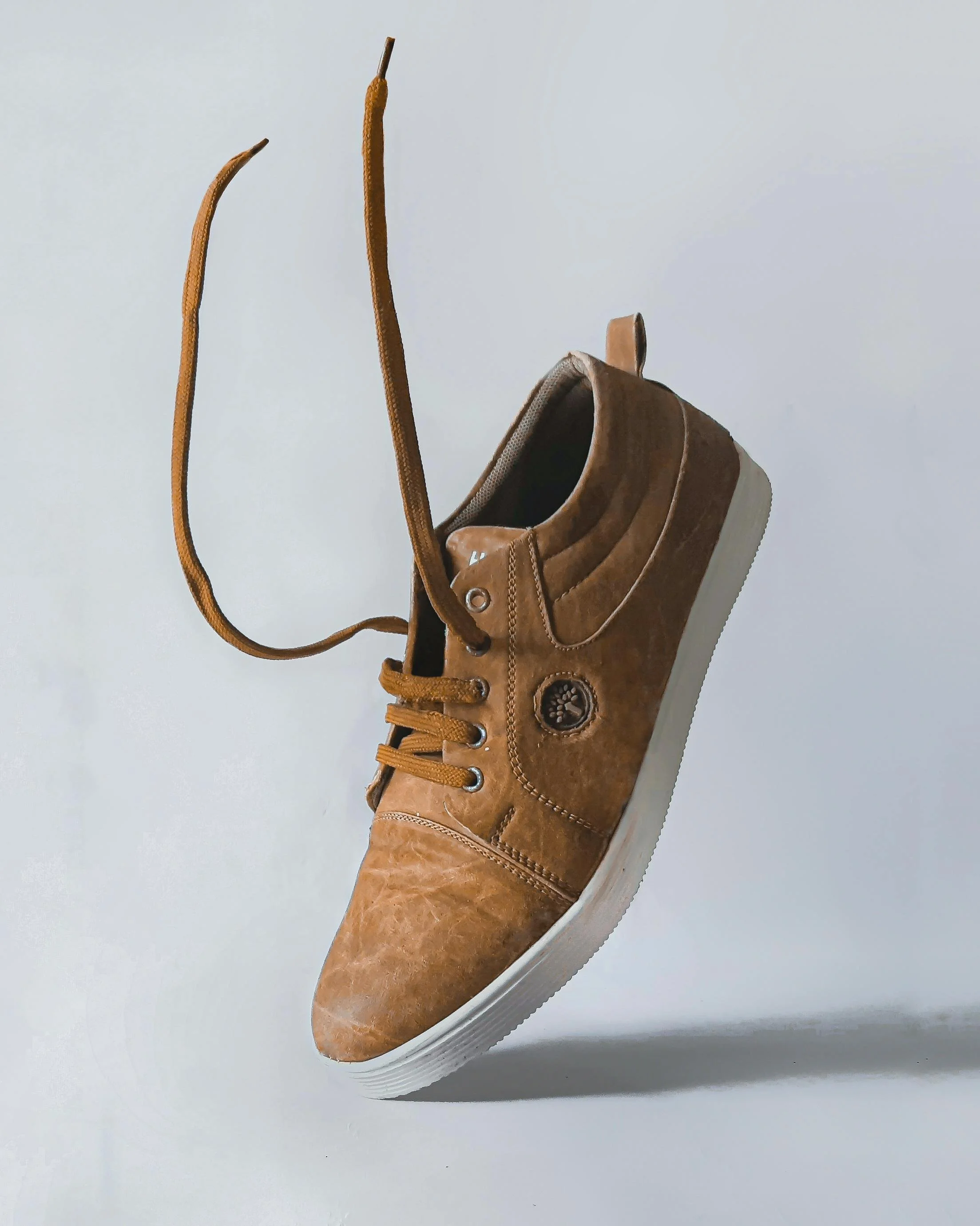 A brown leather sneaker with tan laces, white sole, and a circular logo on the side, tilted to the side against a white background.