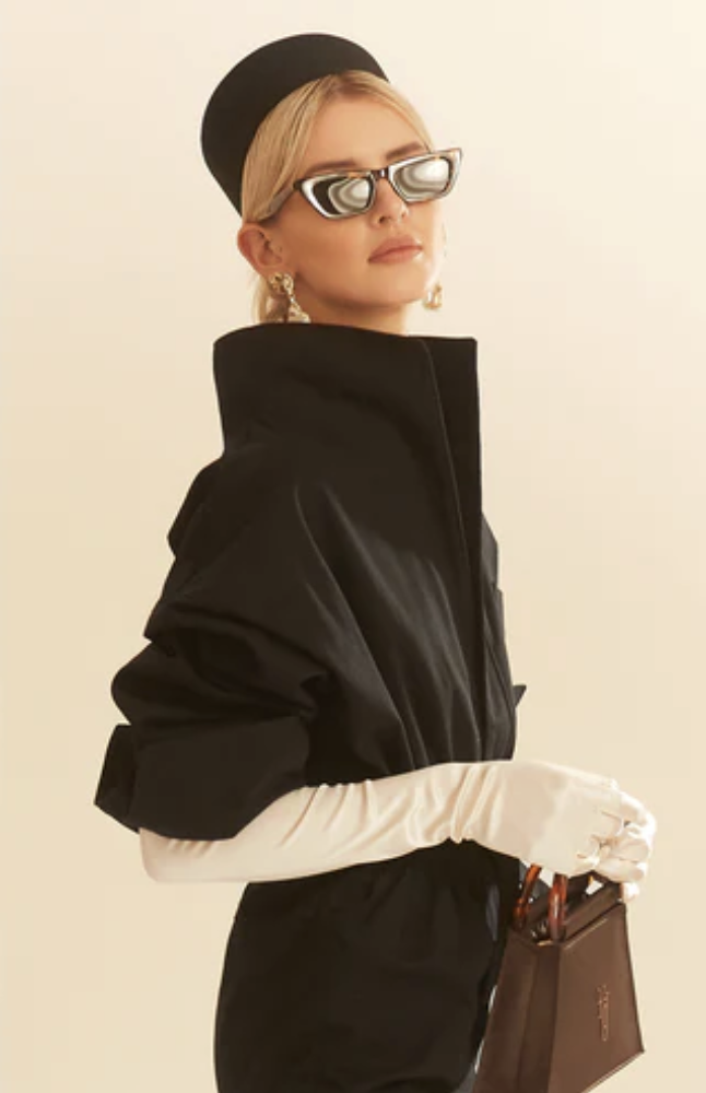 A woman with blonde hair, wearing a black beret, sunglasses, and a black high-collared jacket, holding a brown bag and wearing white gloves.