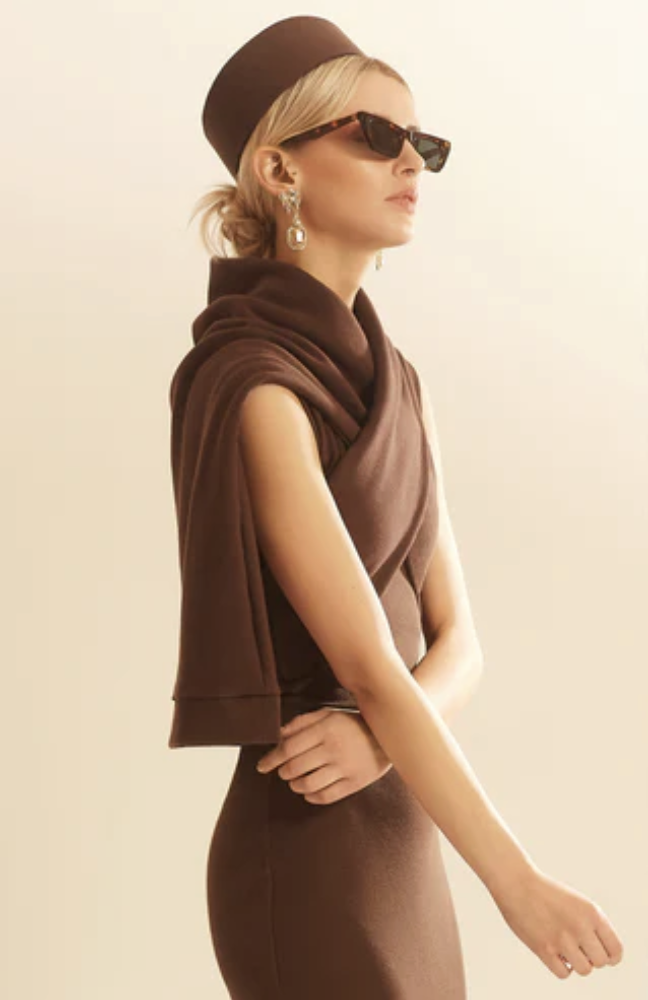 Woman in brown hat and sunglasses, wearing jewelry, wrapped in a brown shawl, with arms crossed, against a neutral background.