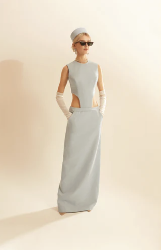 Woman wearing a long, sleeveless, light gray dress with cutouts at the waist, accessorized with gloves, sunglasses, earrings, a hat, and a necklace, standing against a plain background.