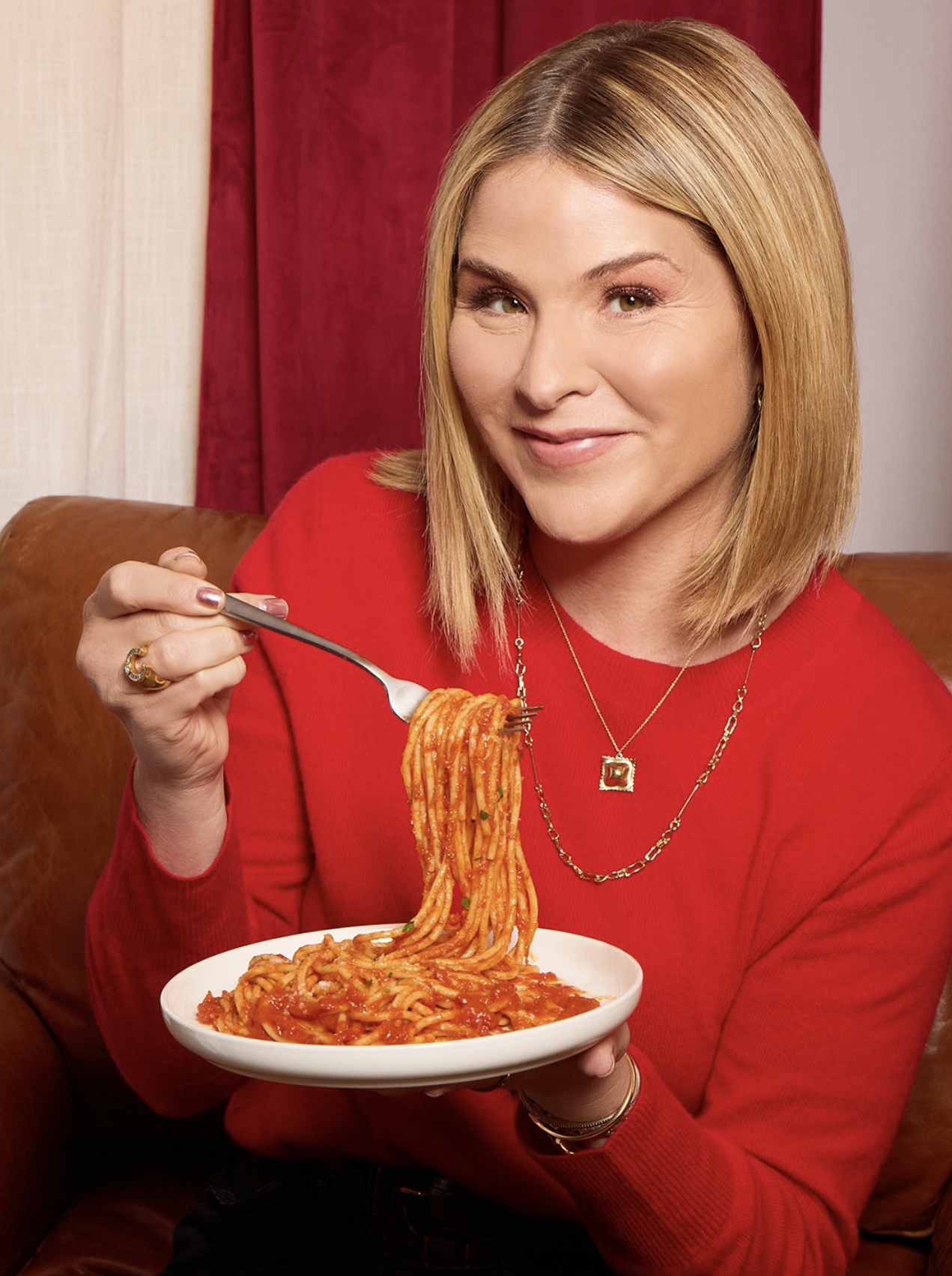 A woman with blonde hair wearing a red sweater, sitting on a brown leather couch, holding a fork with spaghetti on it and smiling, with a plate of spaghetti in her other hand.