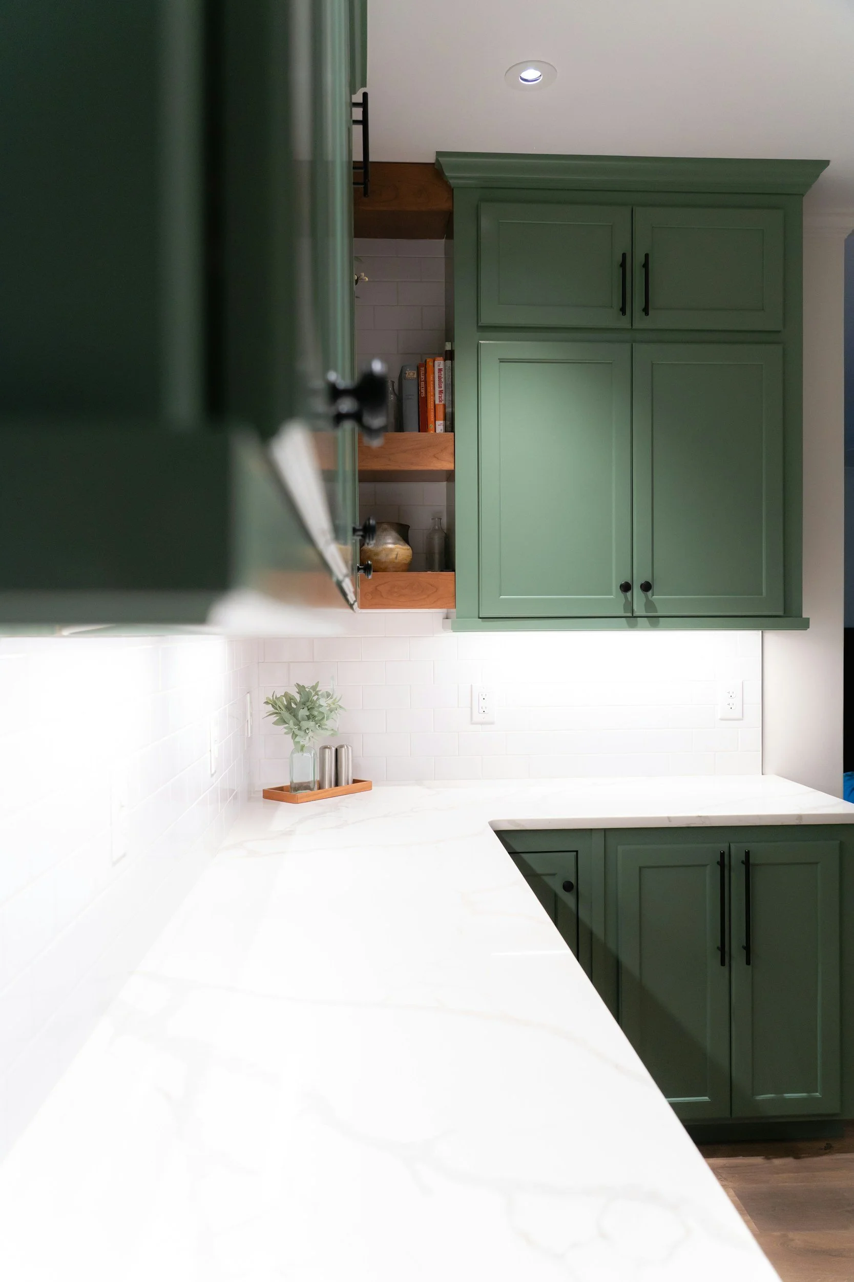 A kitchen with green cabinets, white marble countertops, and white subway tile backsplash. An open shelf with cookbooks and jars is visible, along with a small plant and salt and pepper shakers on the counter.