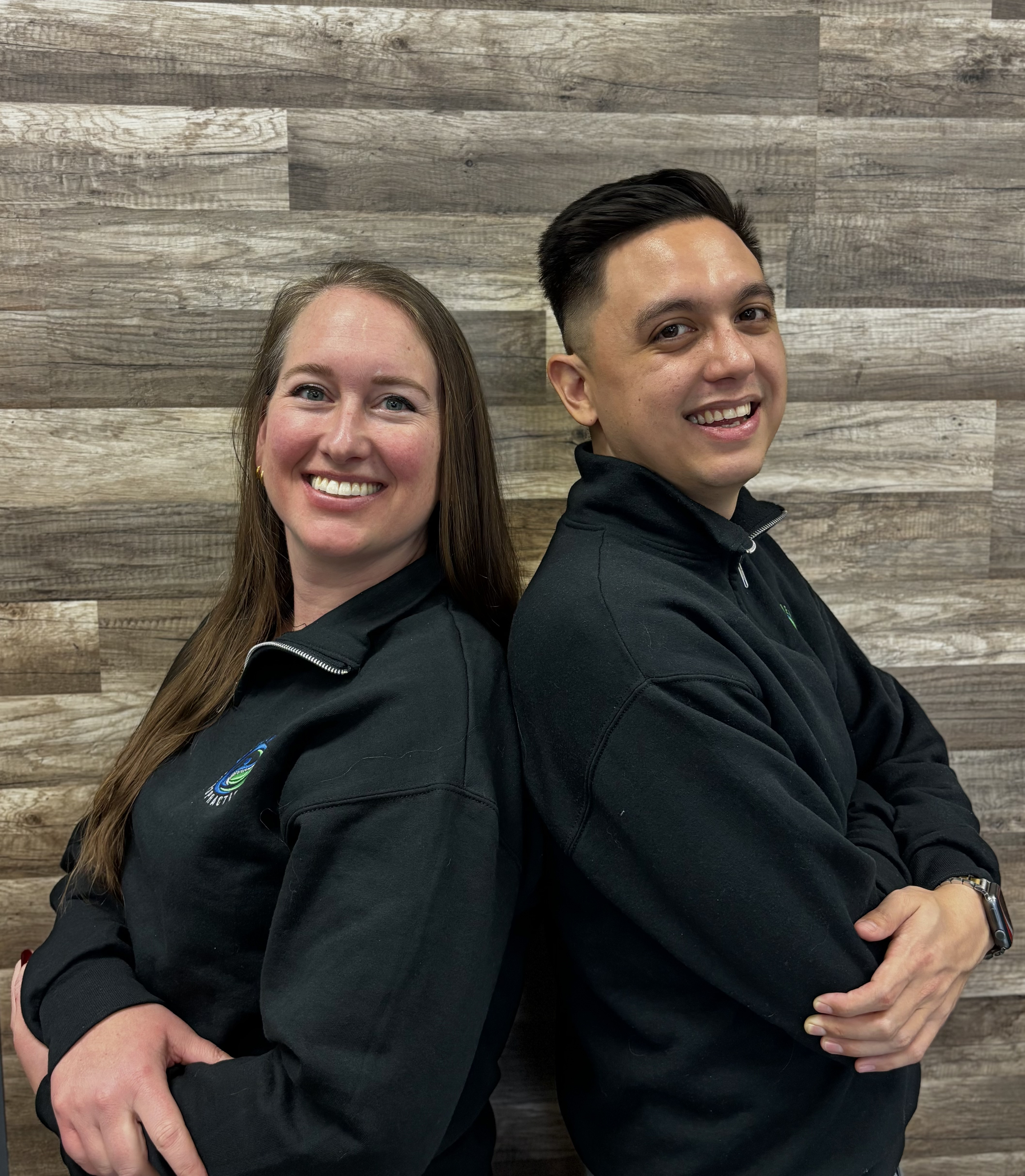 Two smiling young adults, a woman with long brown hair and a man with short dark hair, standing back-to-back with arms crossed in front of a wooden wall, wearing black zip-up jackets.