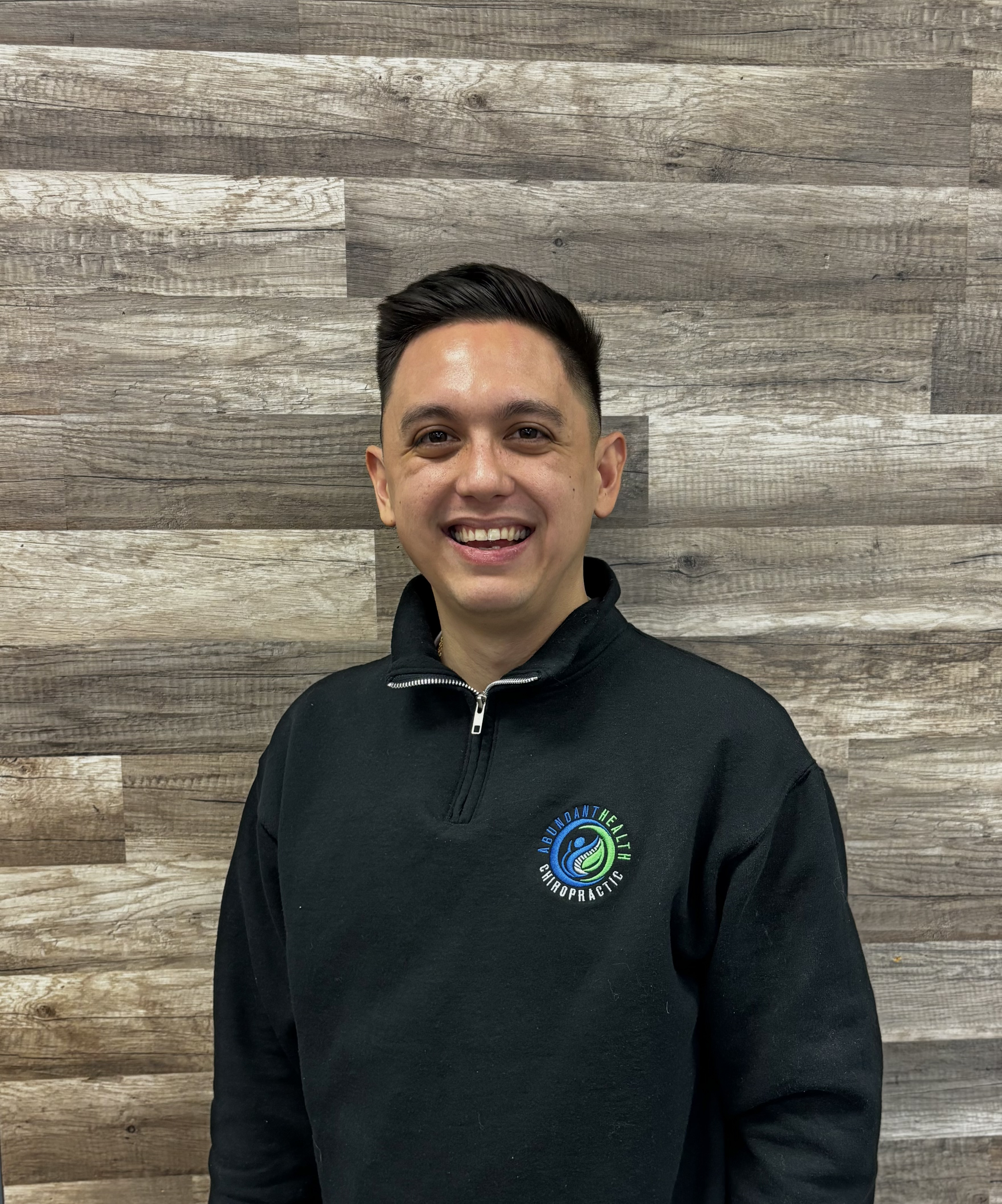 A young man with short dark hair smiling and standing in front of a wooden wall, wearing a black pullover with a logo of 'Abundant Health Chiropratic'.