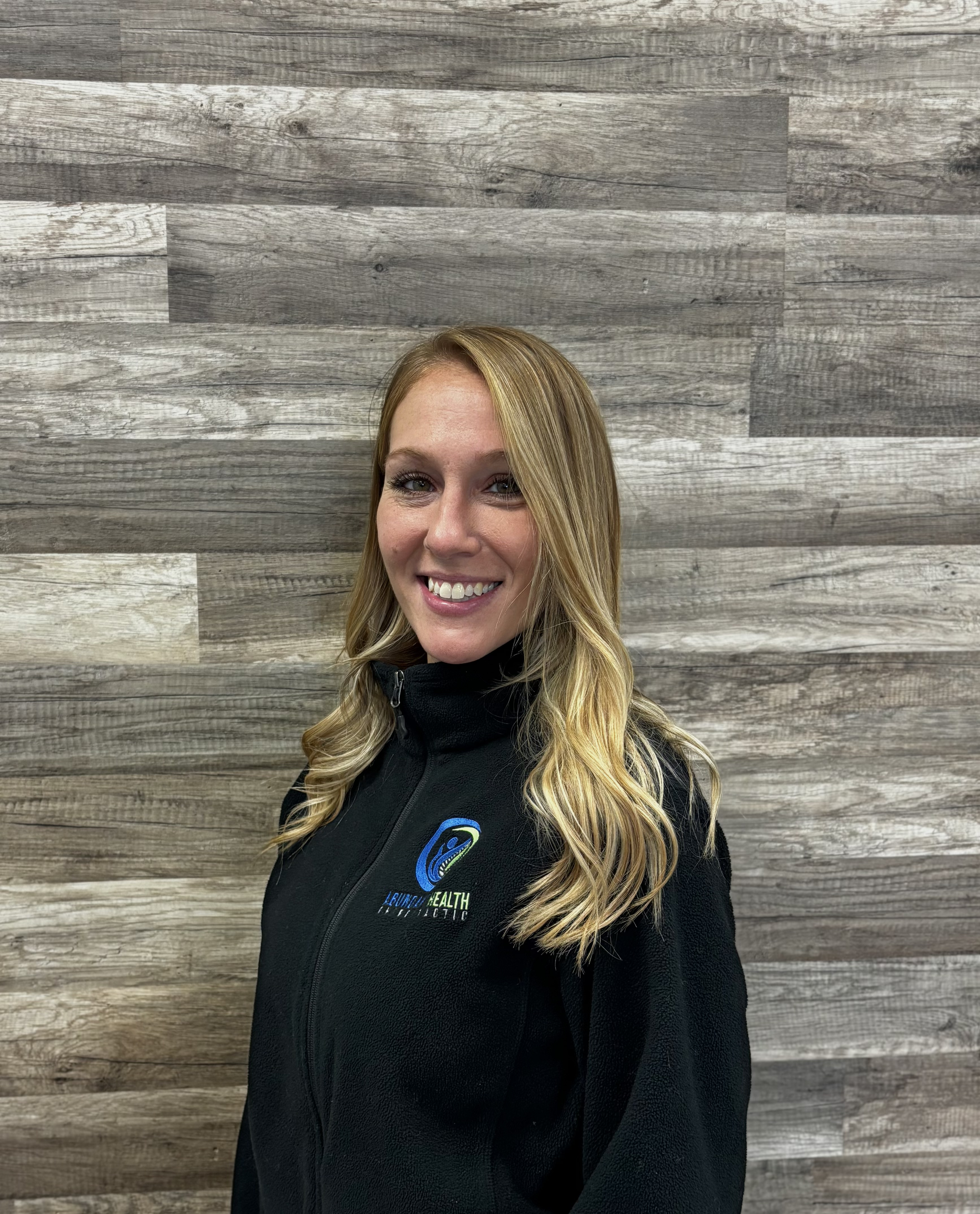 A woman with long blond hair wearing a black jacket with a "Source of Health Chiropractic" logo, standing in front of a wooden wall.