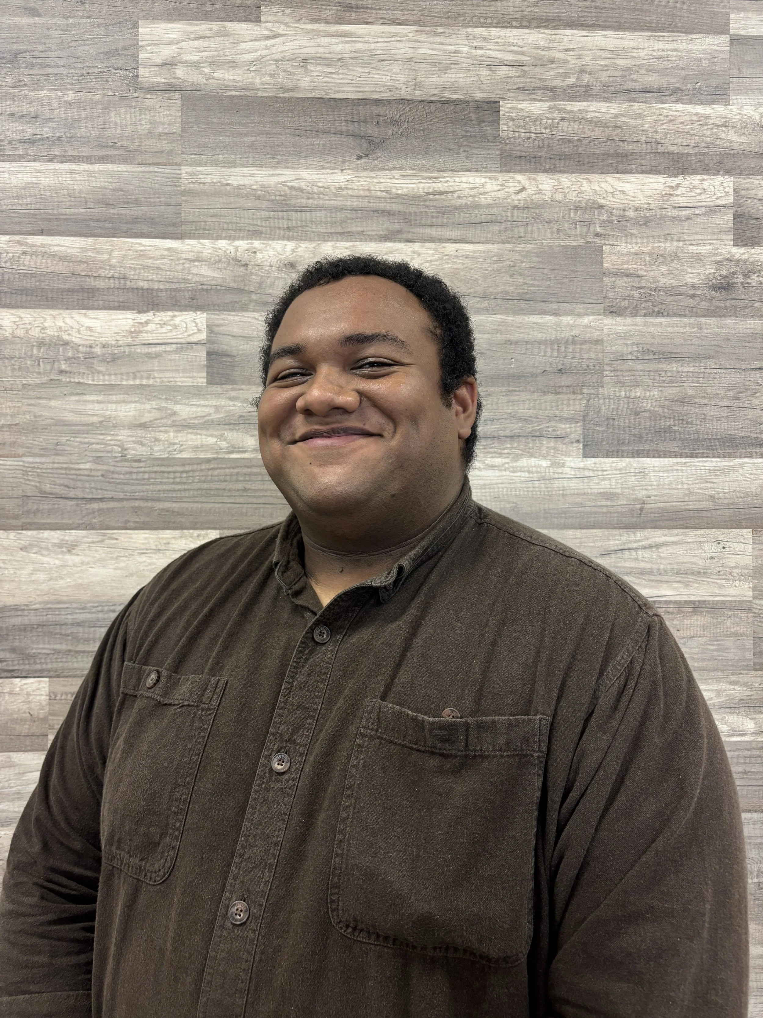 Close-up of a smiling man with short curly black hair, wearing a brown button-up shirt, standing against a wood-paneled wall.