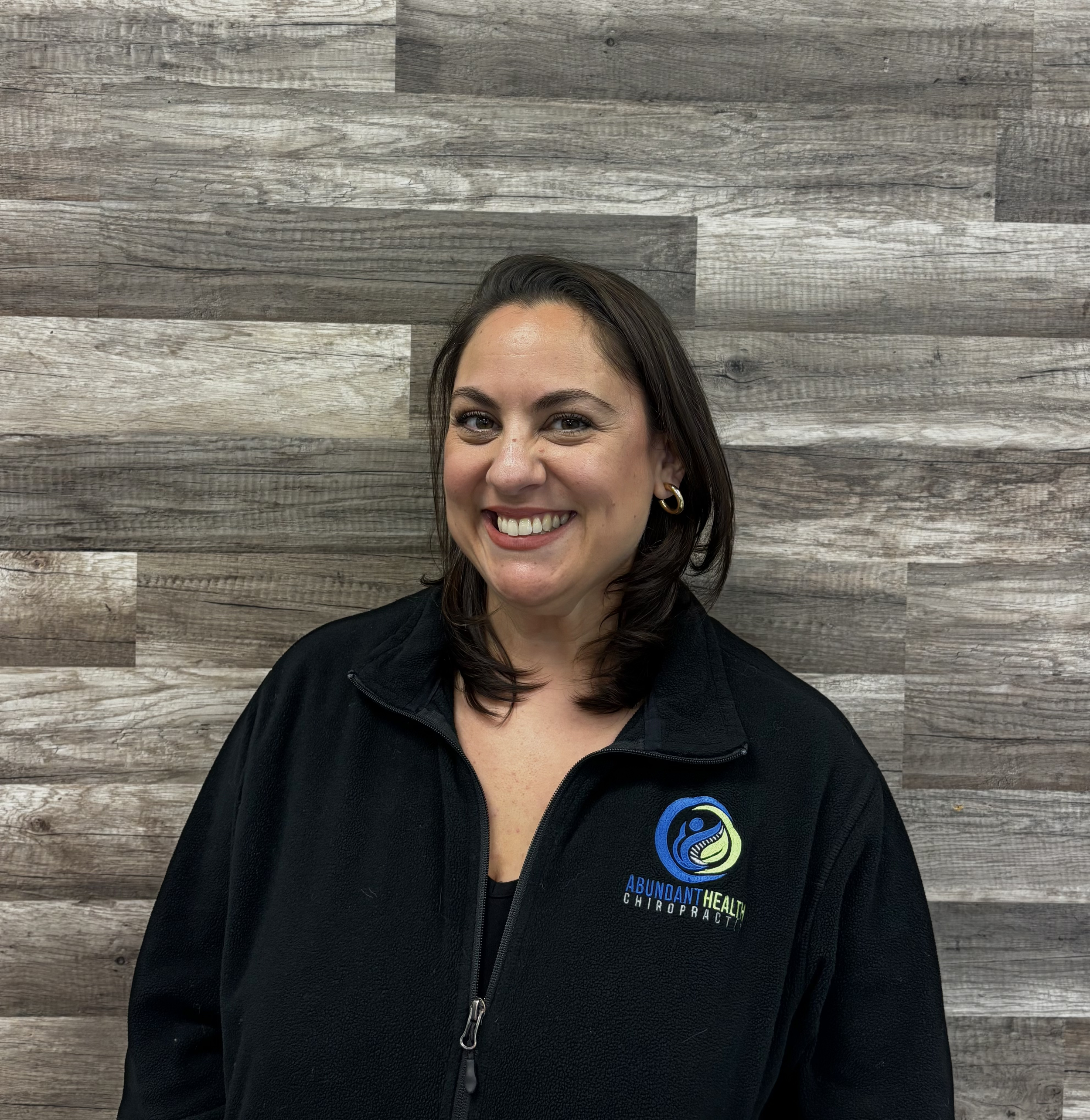 Woman with shoulder-length dark hair smiling, wearing a black fleece jacket with a logo for Abundant Health Chiropractic, standing in front of a wooden wall.