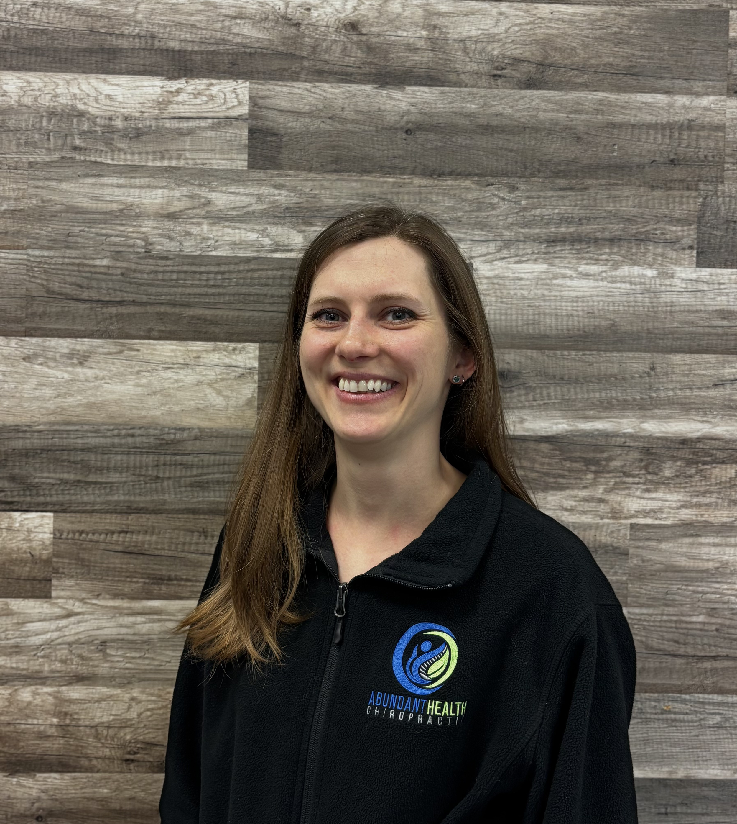 Portrait of a smiling woman with long brown hair wearing a black zip-up jacket with 'Abundant Health Chiropractic' logo, standing against a wooden panel background.