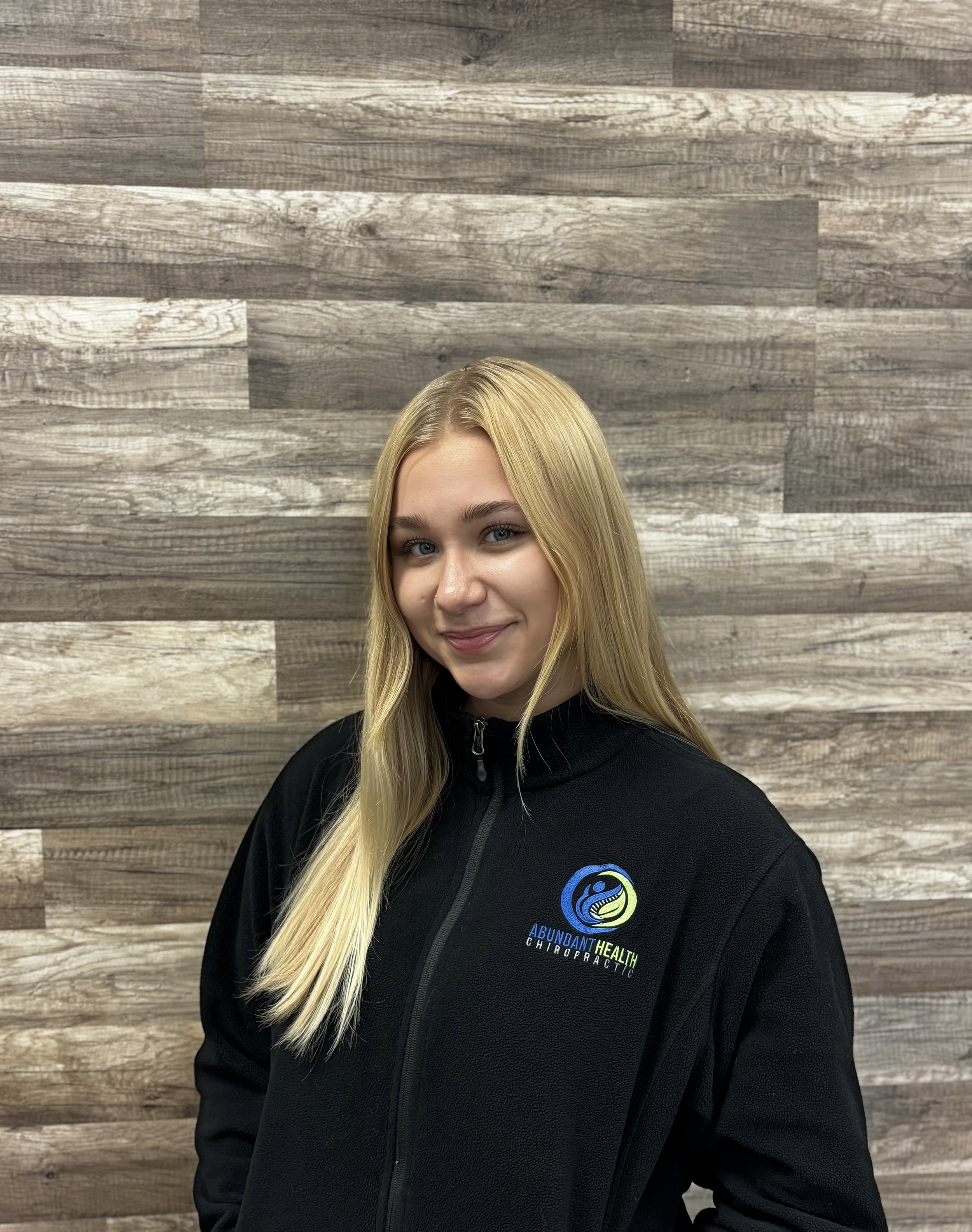 A young woman with long blonde hair smiling, wearing a black zip-up jacket with a logo that reads 'Abundant Health Chiropractic' in front of a wooden-textured wall.