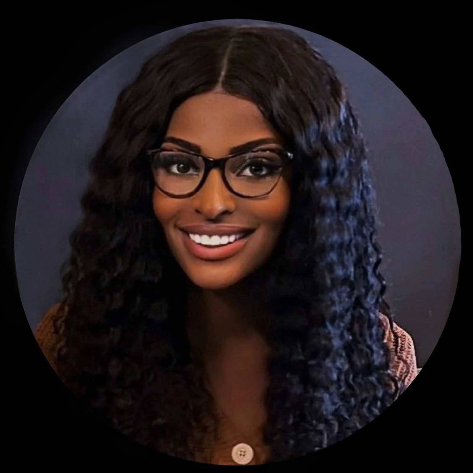 A young woman with long, curly dark hair and glasses, smiling at the camera against a dark background.