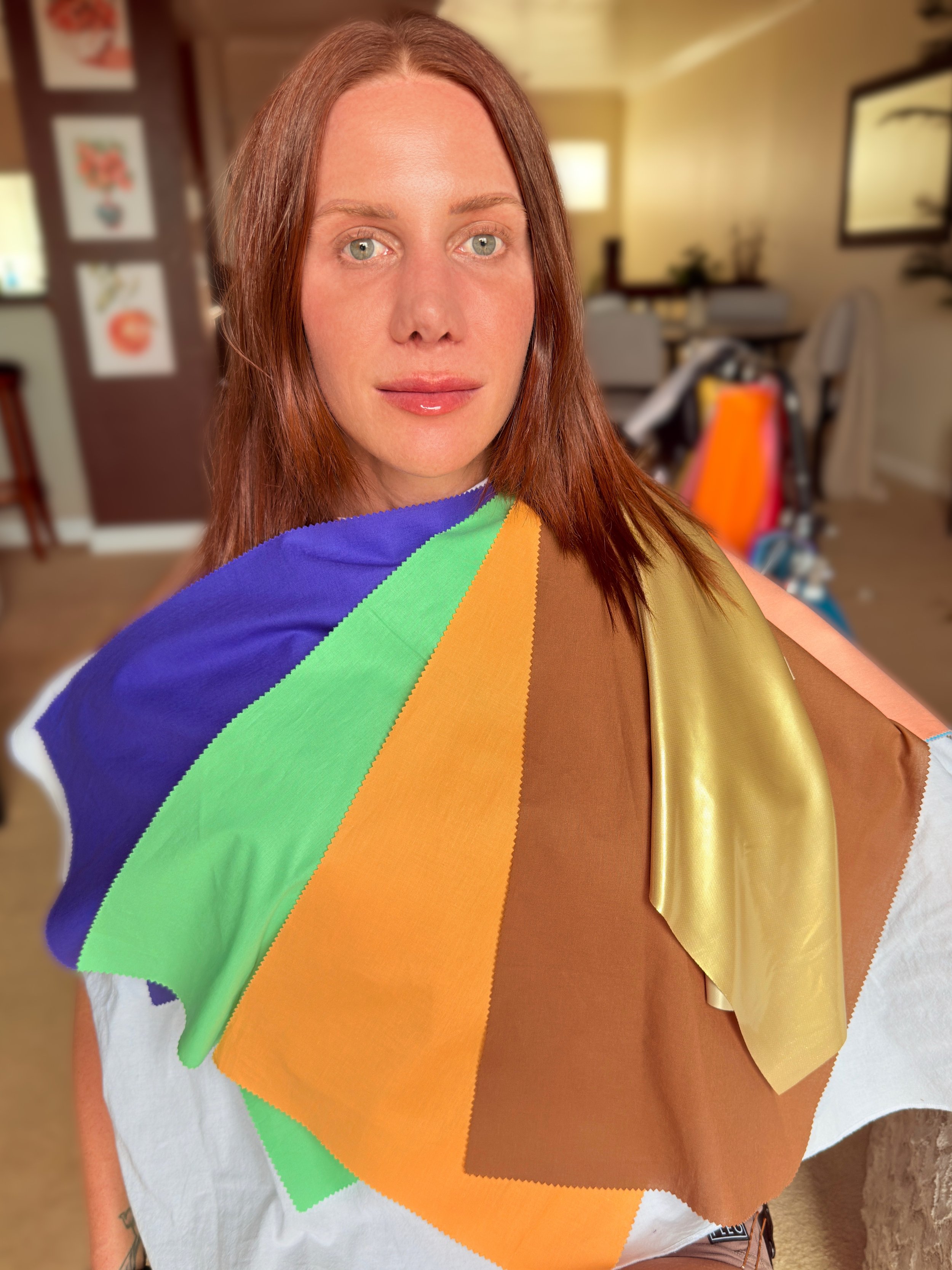A woman with red hair and light skin looking at the camera, wearing a rainbow-colored color analysis drapes over a white shirt, in a cozy room with framed pictures and a dining area in the background.