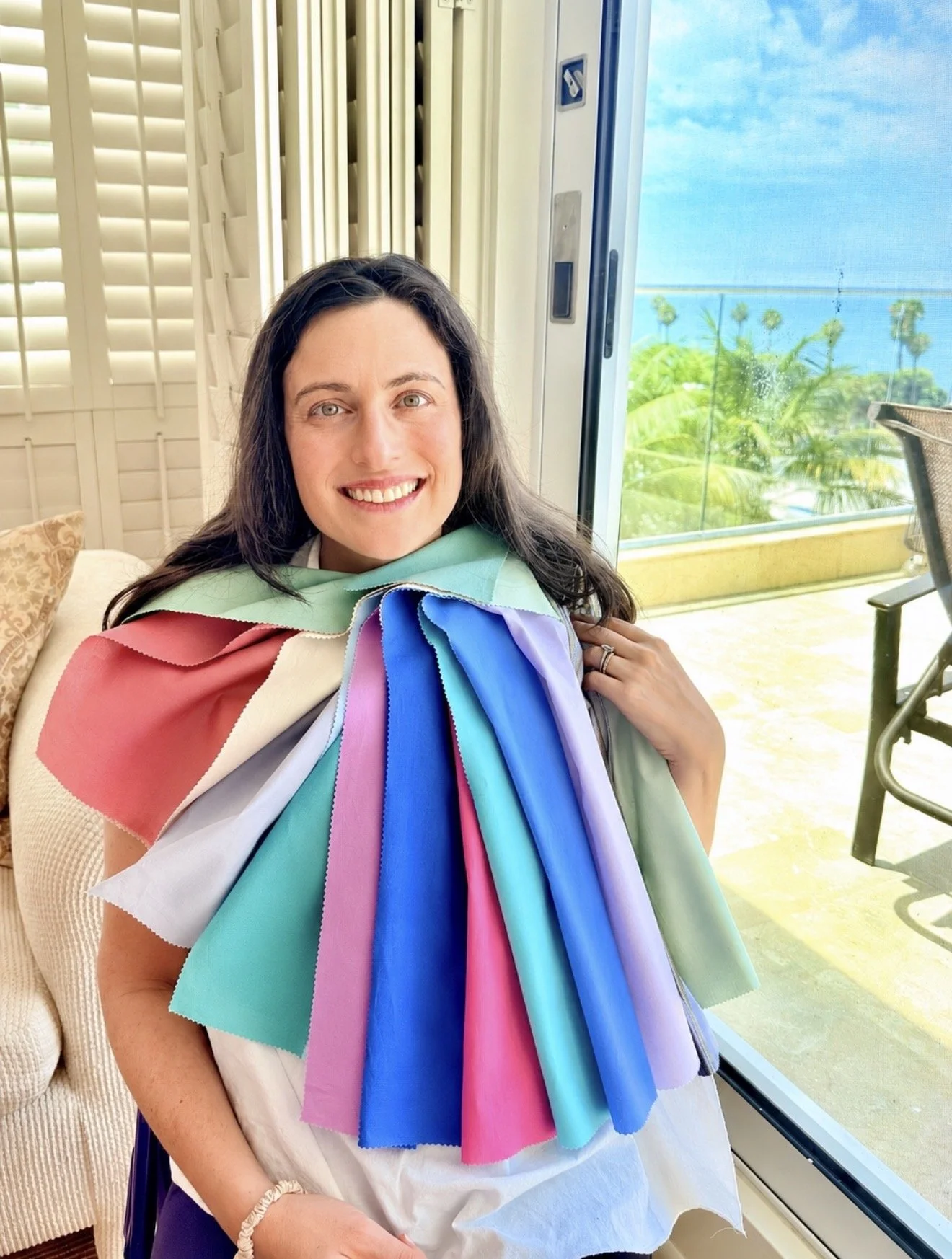 A woman sitting indoors near a glass door with a view of greenery and the ocean. She has long dark hair and is smiling. She is wearing fabric samples in various colors draped over her shoulders after her color analysis session