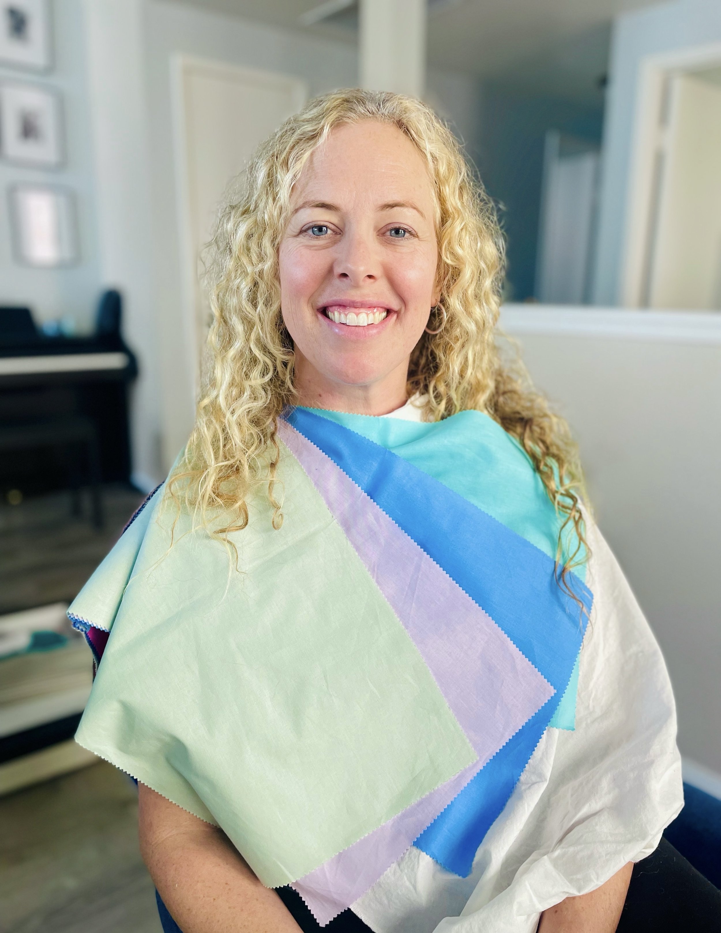 A woman with blonde curly hair smiling at the camera wearing a colorful cloth in a room with a piano and picture frames on the wall.