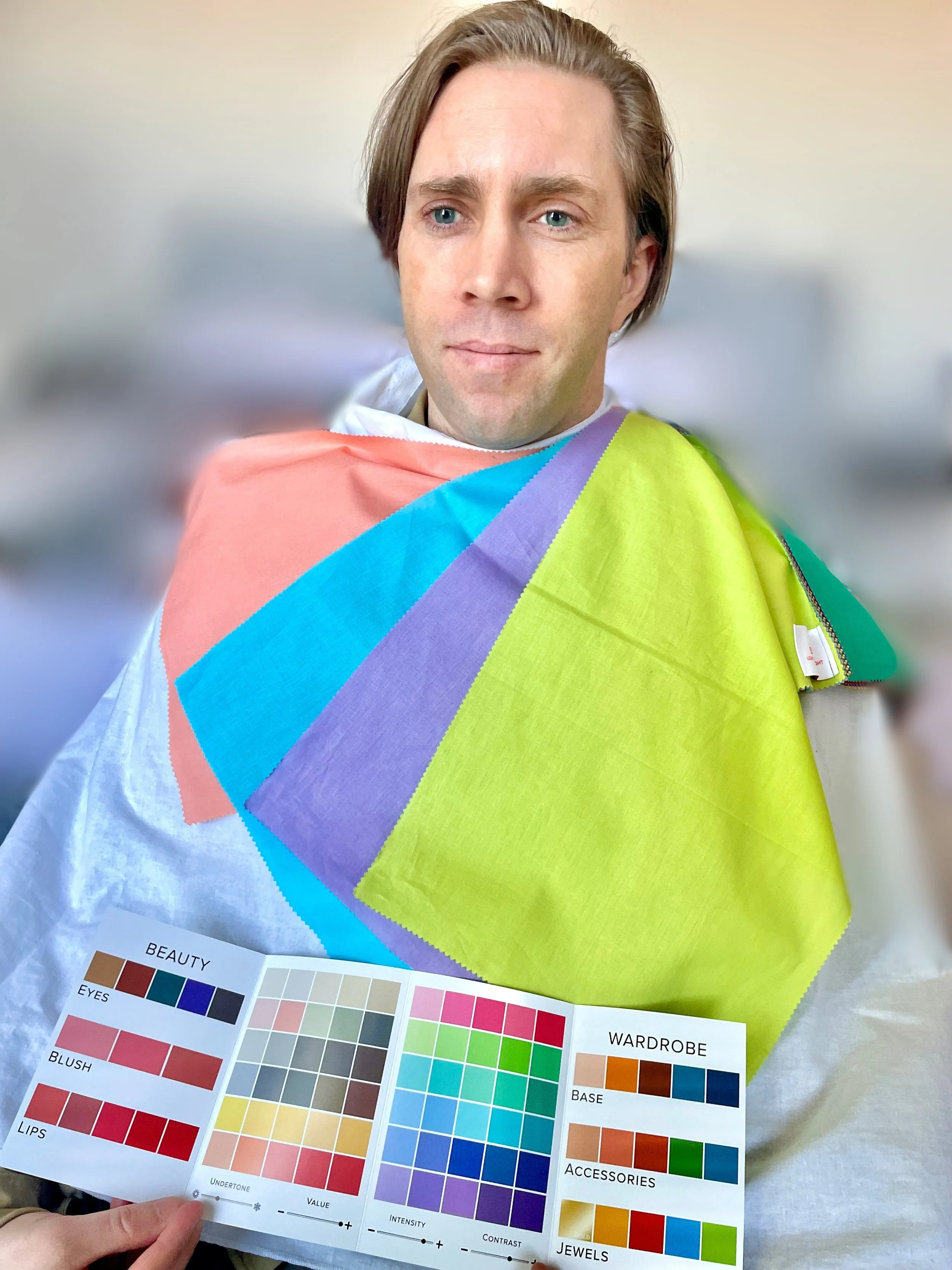 Person with short brown hair and blue eyes wearing a colorful quilted cape, holding a color palette chart.