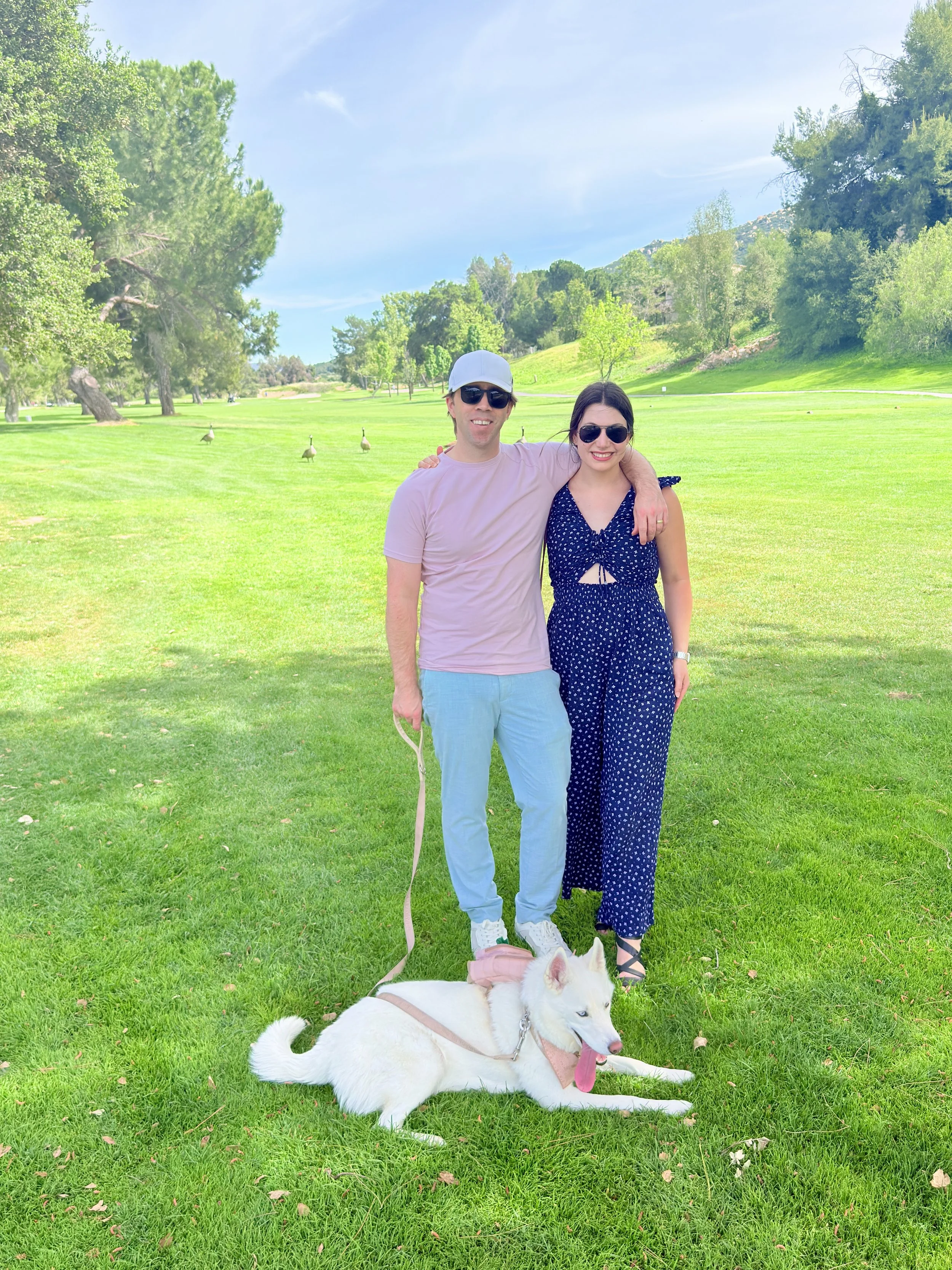 A smiling young man and woman stand on a lush green golf course, with the man wearing sunglasses, a white cap, and pastel-colored casual clothes, and the woman wearing sunglasses and a navy blue dress with white polka dots. They are posing with their arms around each other. A white husky dog with blue eyes, wearing a pink harness and bandana, is lying on the grass in front of them. In the background, there are trees, shrubs, and a clear blue sky.