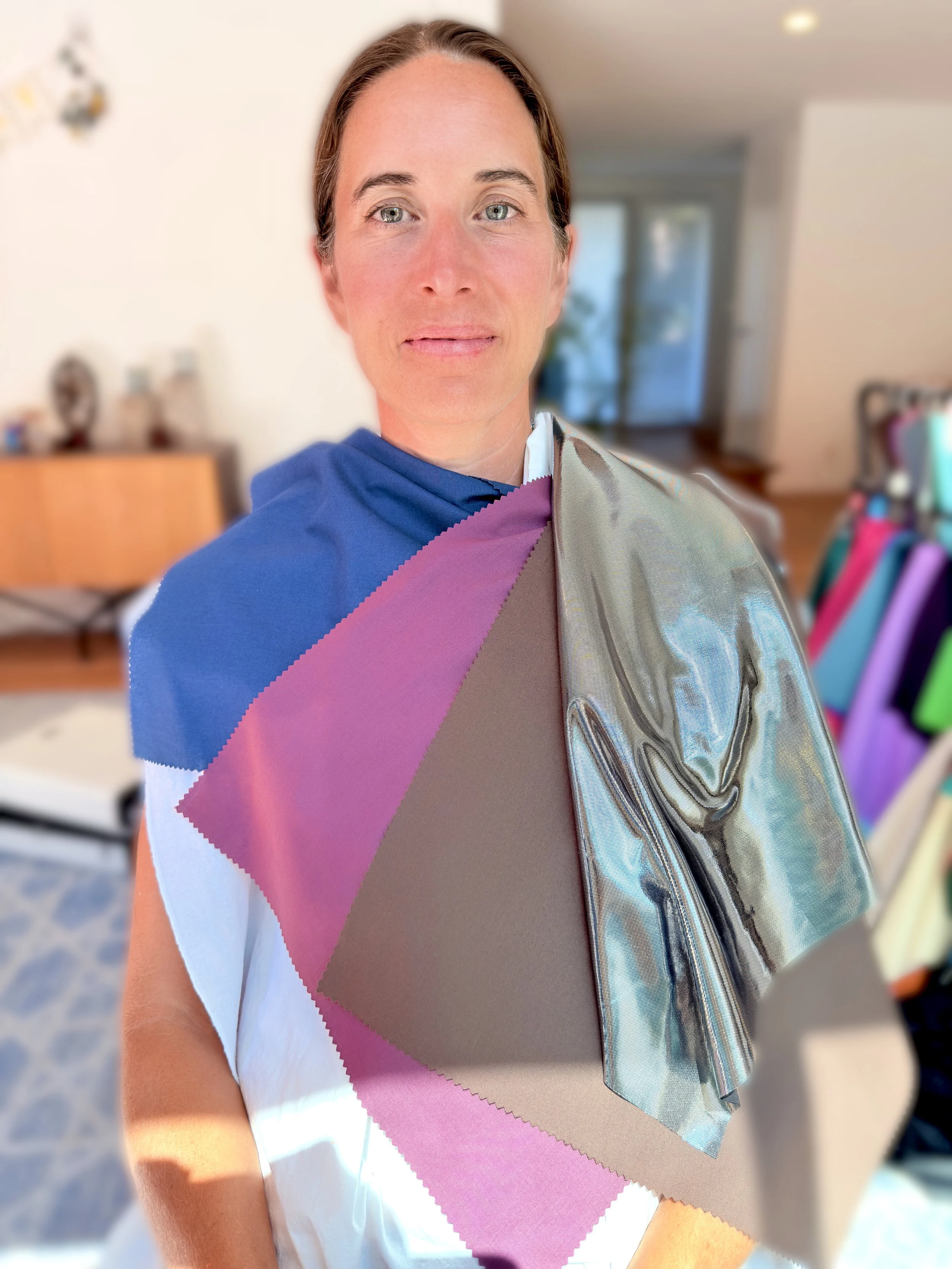 A woman with short brown hair and blue eyes standing indoors, wearing a white shirt and color analysis drapes over her shoulder. The background shows a living room with a blurred piece of furniture and a colorful display on the right side.