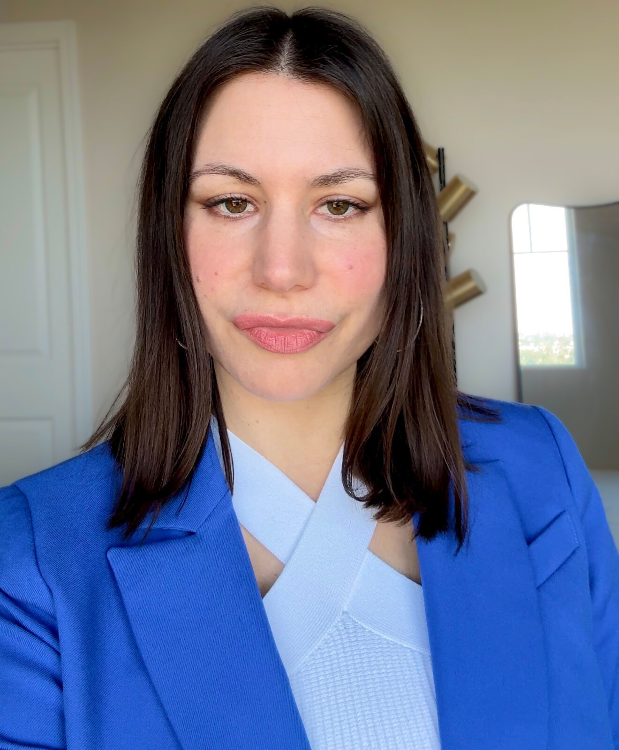 A woman with brown hair and light makeup wearing a blue blazer and a white top, standing indoors with a mirror and window in the background.